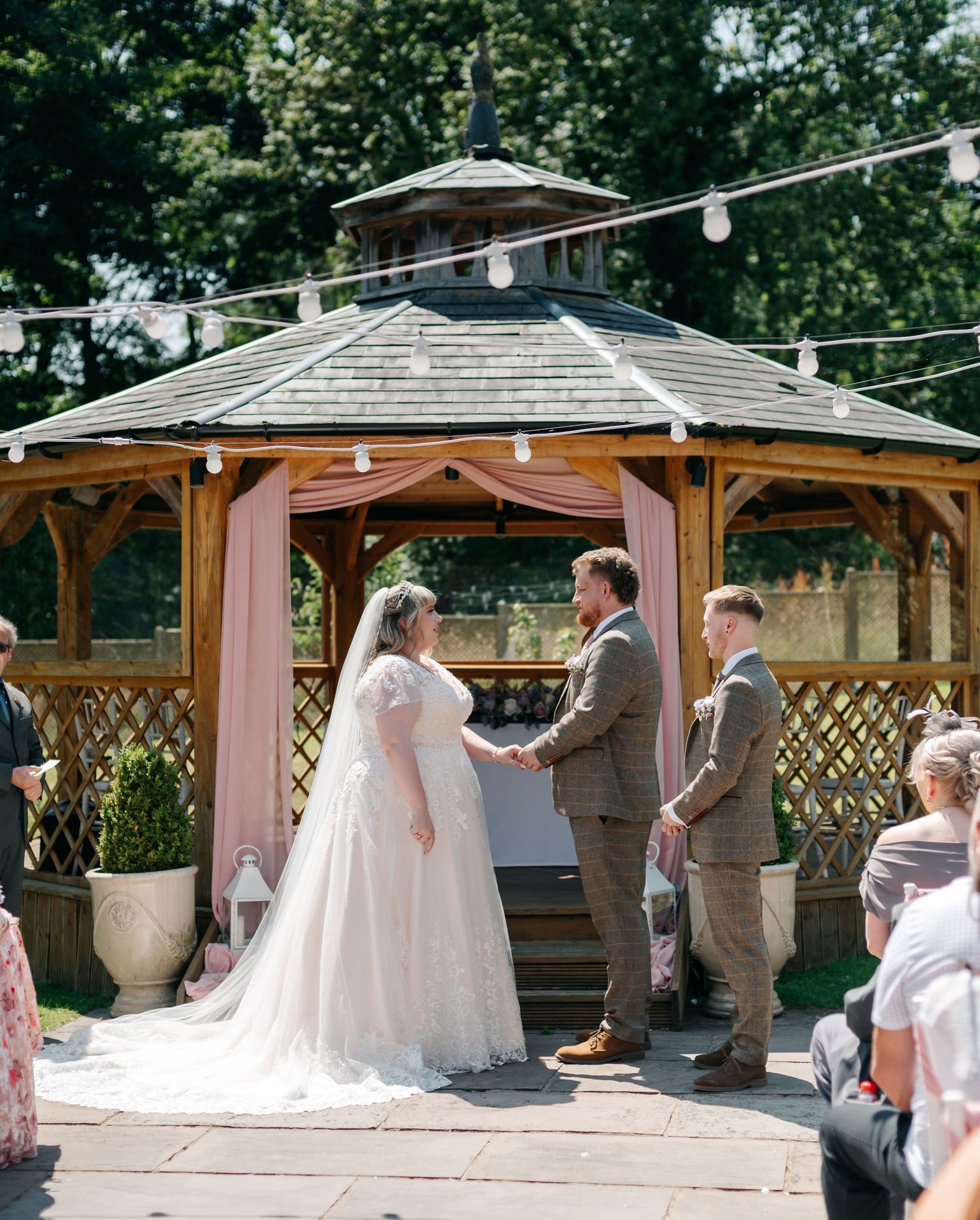 Bride and groom exchange vows at romantic outdoor gazebo ceremony decorated with pink draping and fairy lights.