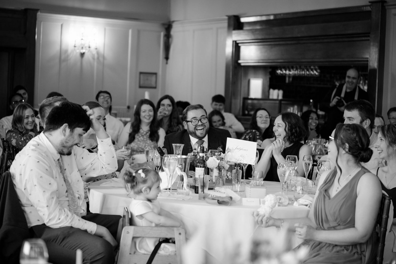 Black and white photo of smiling wedding guests laughing at a reception table with a child, glasses, and a small sign.