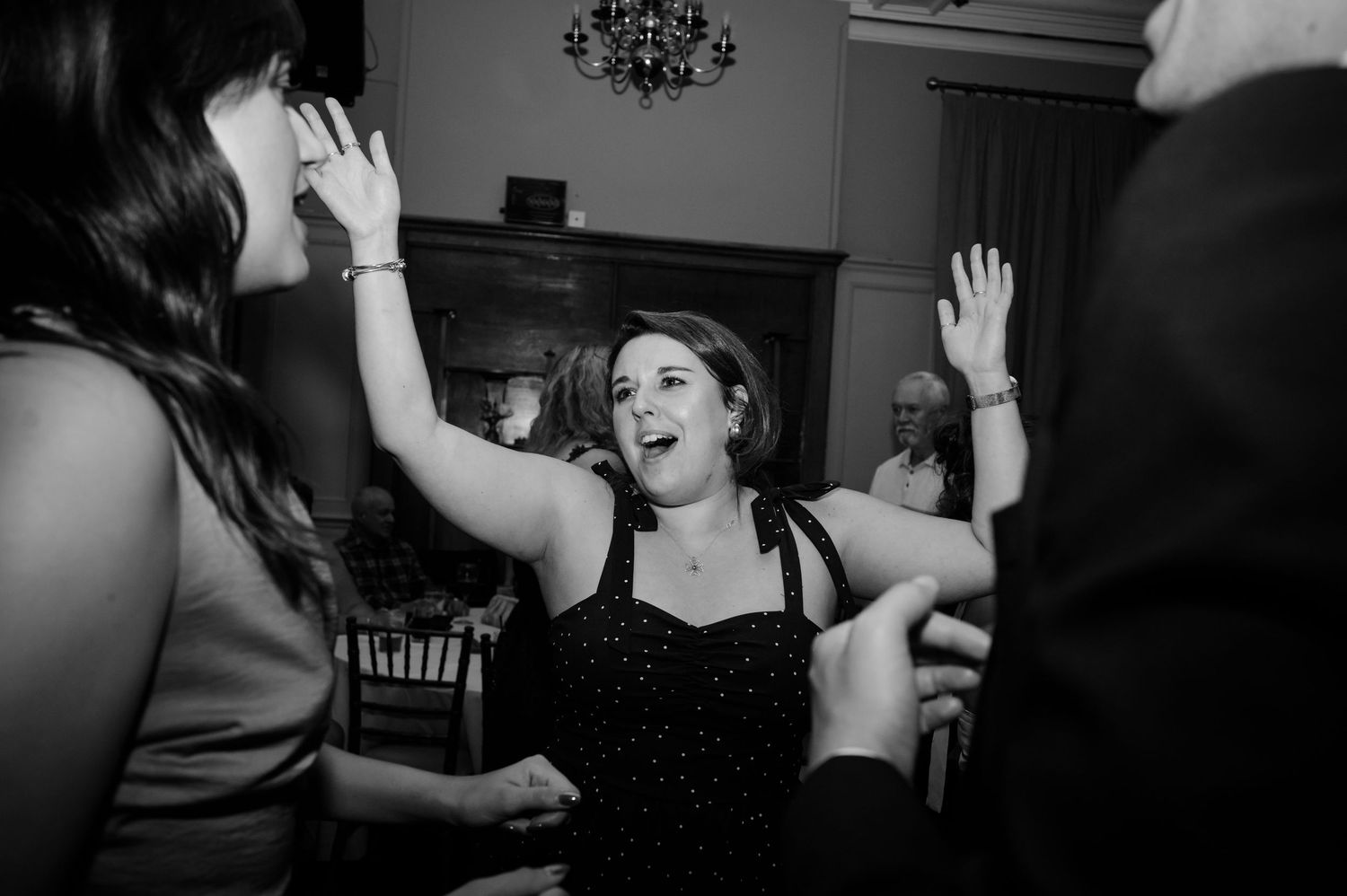 Black and white photo of a joyful woman in a sparkly top dancing with arms raised among guests at a lively indoor wedding reception.