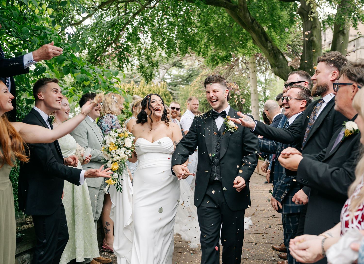 Smiling bride in a white gown and groom in a dark suit hold hands as wedding guests cheer and throw confetti along a leafy outdoor path.