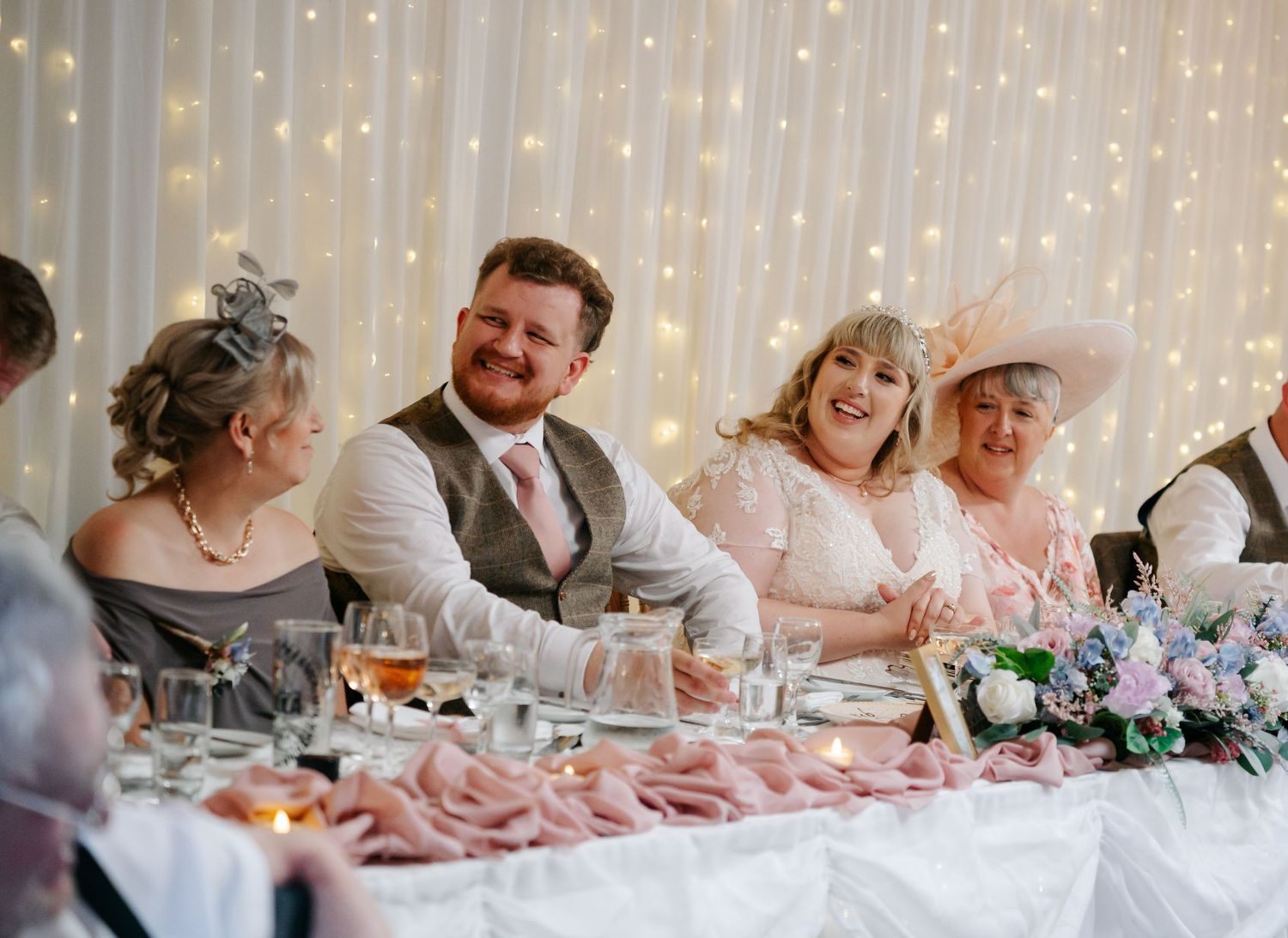 Happy wedding party guests laughing at a decorated head table with fairy lights backdrop.