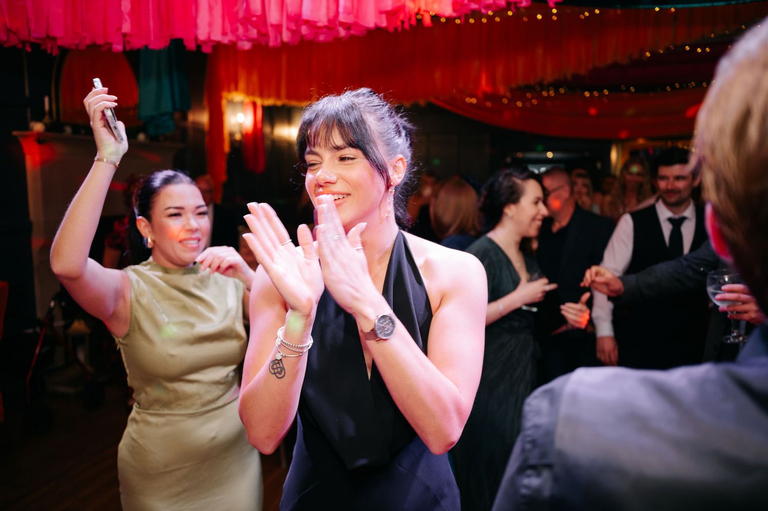 Wedding guests dance and celebrate on a dance floor under red mood lighting and decorative string lights.