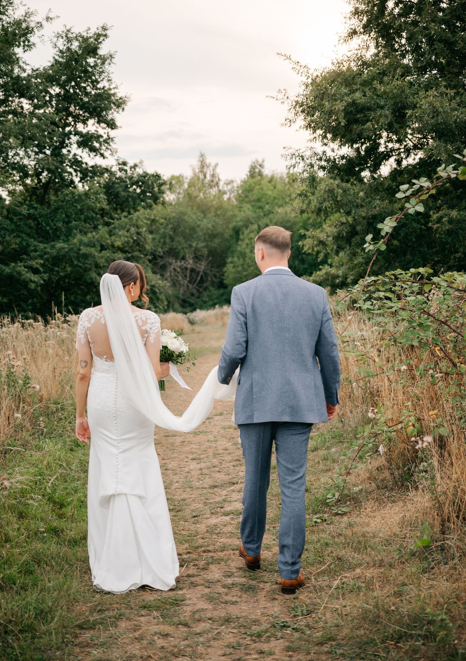 Newlyweds walking hand in hand down a rustic path surrounded by greenery.