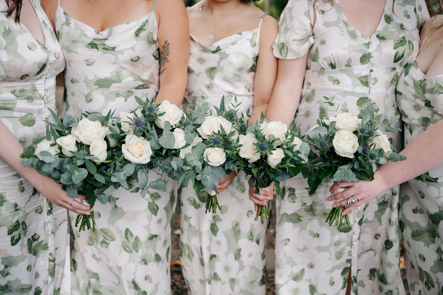 Bridesmaids in floral print dresses hold elegant white rose bouquets with greenery against a soft background.
