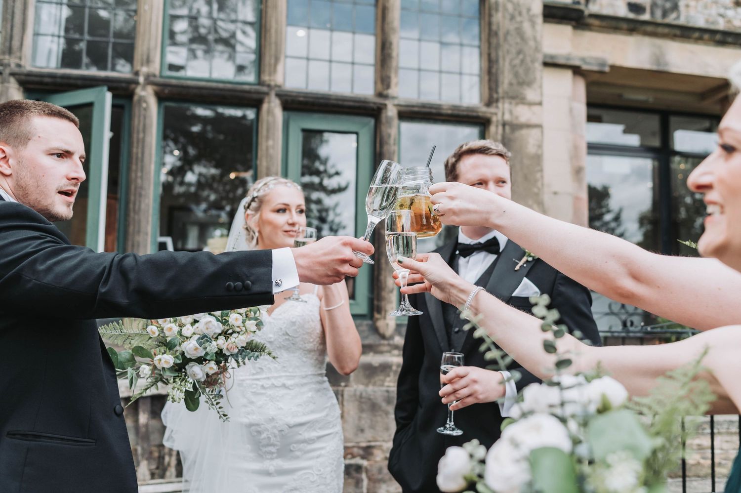 Wedding party toasts with champagne glasses outside historic stone building.