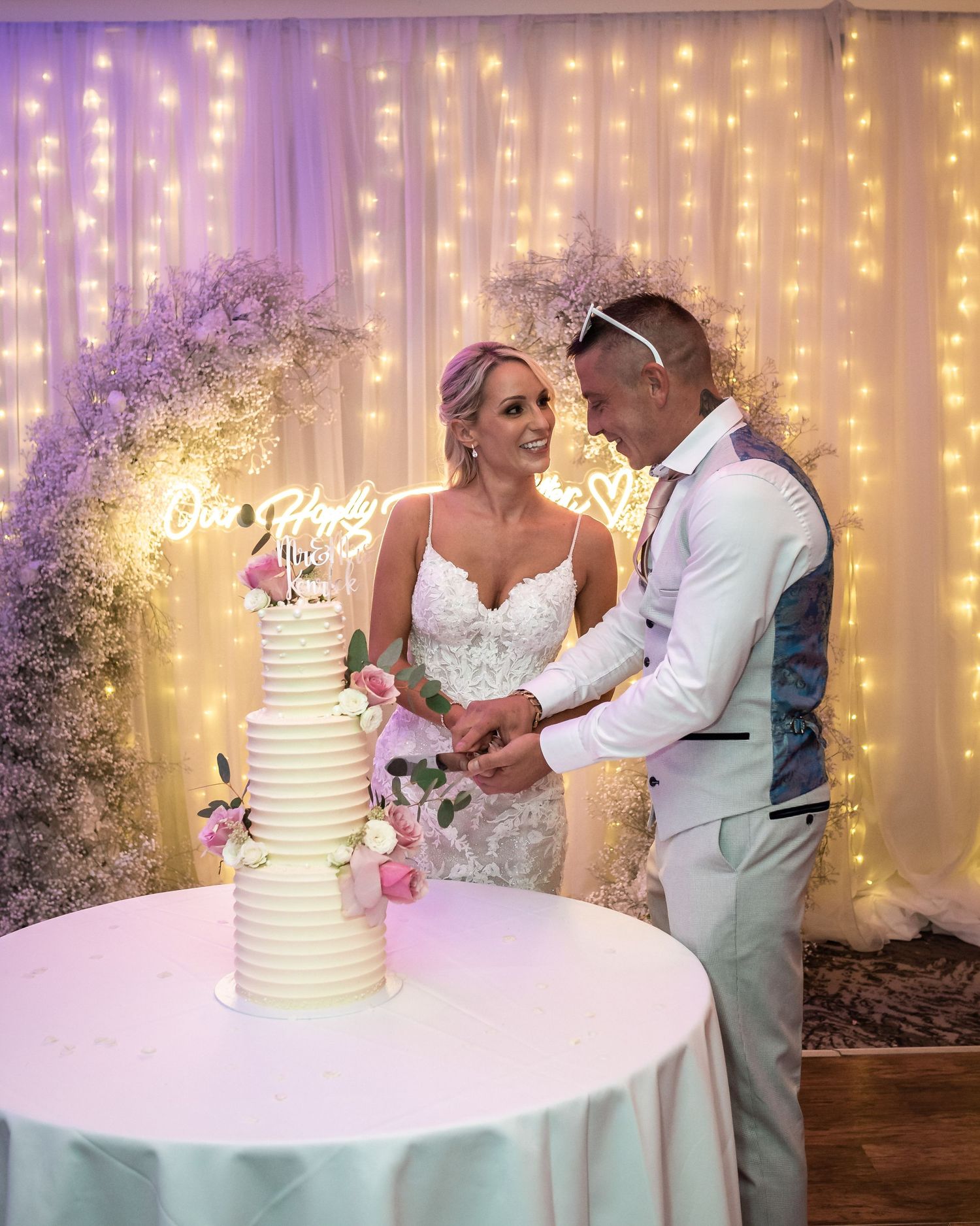 Newlyweds cutting their wedding cake against a backdrop of twinkling lights and purple floral arch.