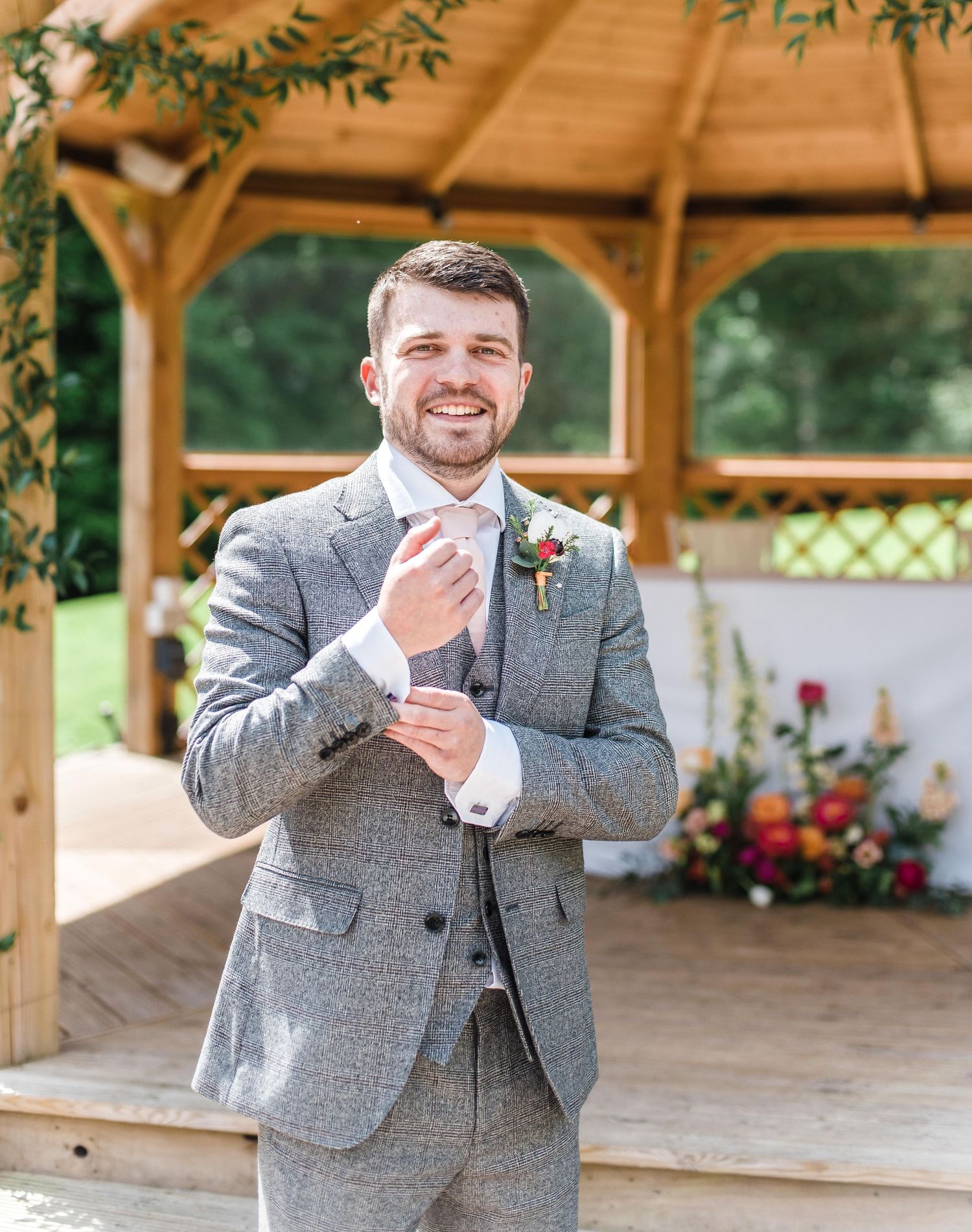 Groom in grey tweed suit adjusts tie while standing in wooden gazebo with floral arrangements.