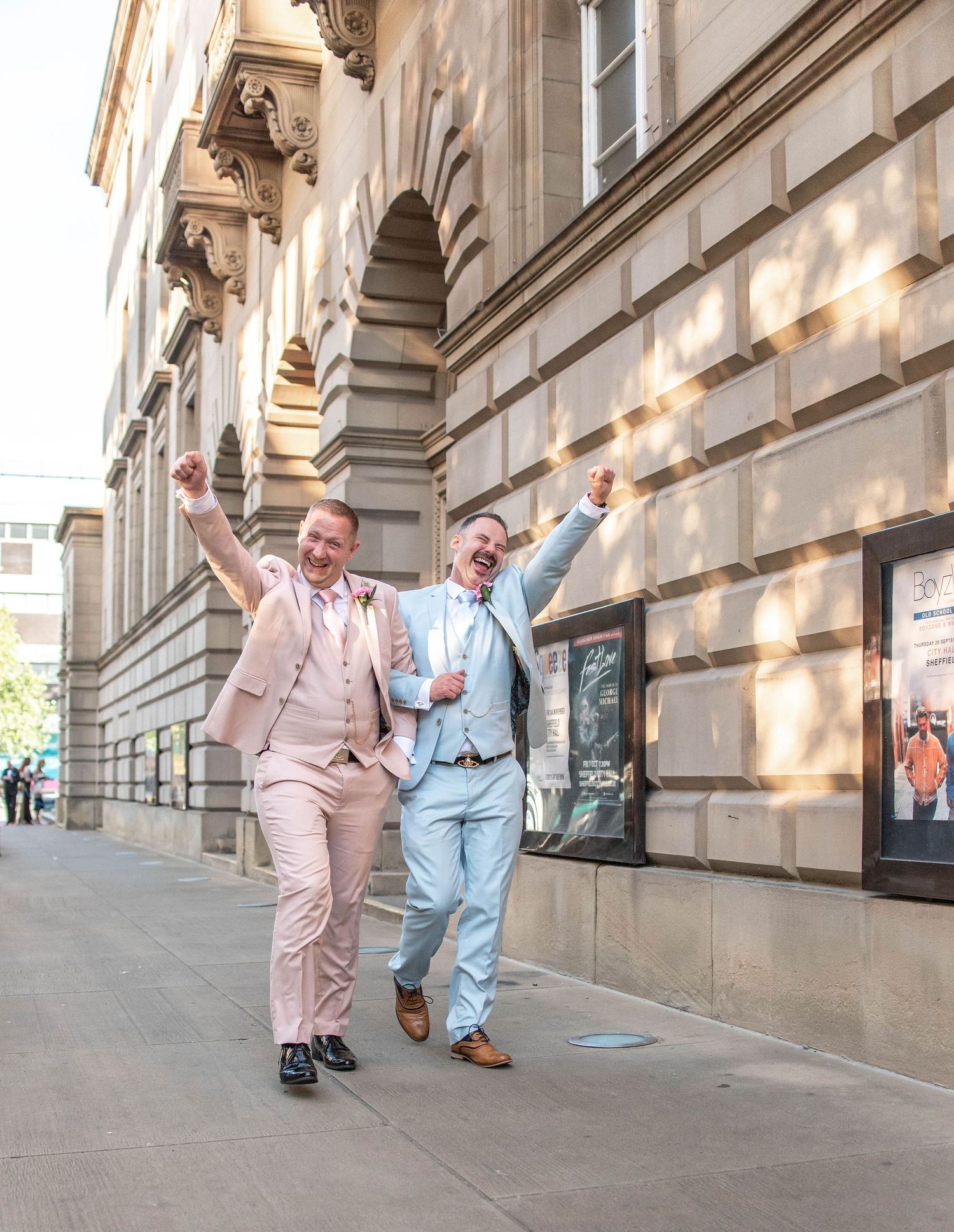 Two people in pastel suits joyfully walking and celebrating outside a building.