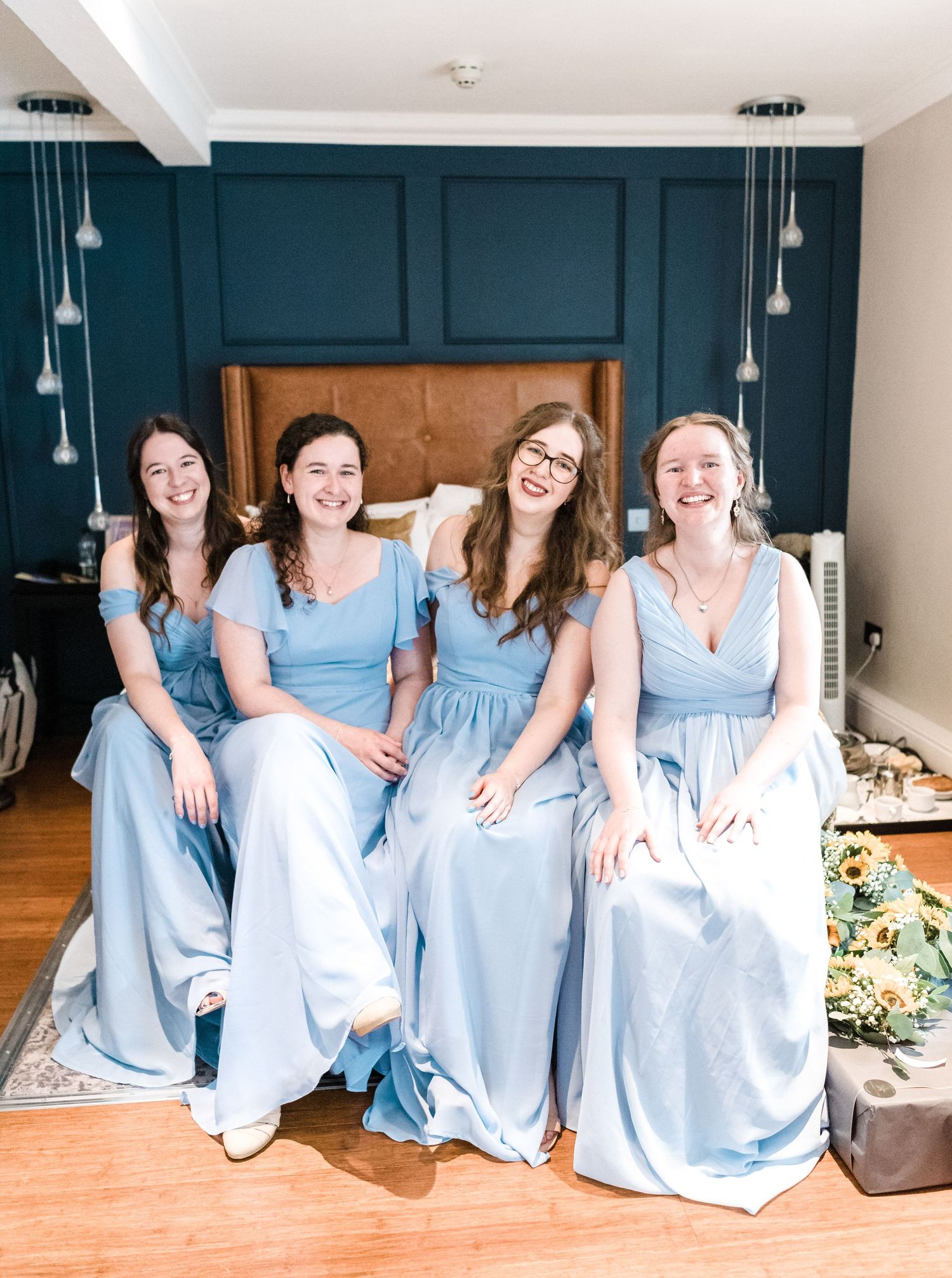Group wearing matching light blue dresses sit together on furniture against a dark teal wall with pendant lights.
