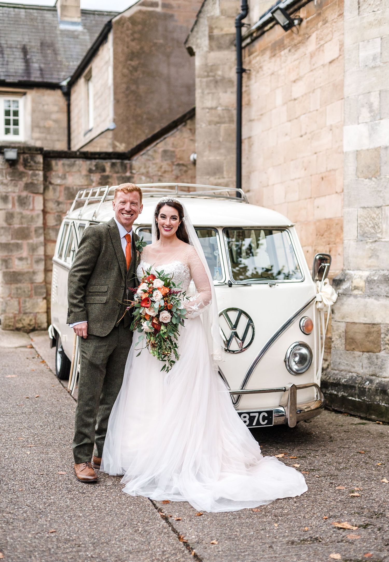 Wedding couple poses with classic white Volkswagen van outside historic stone building.