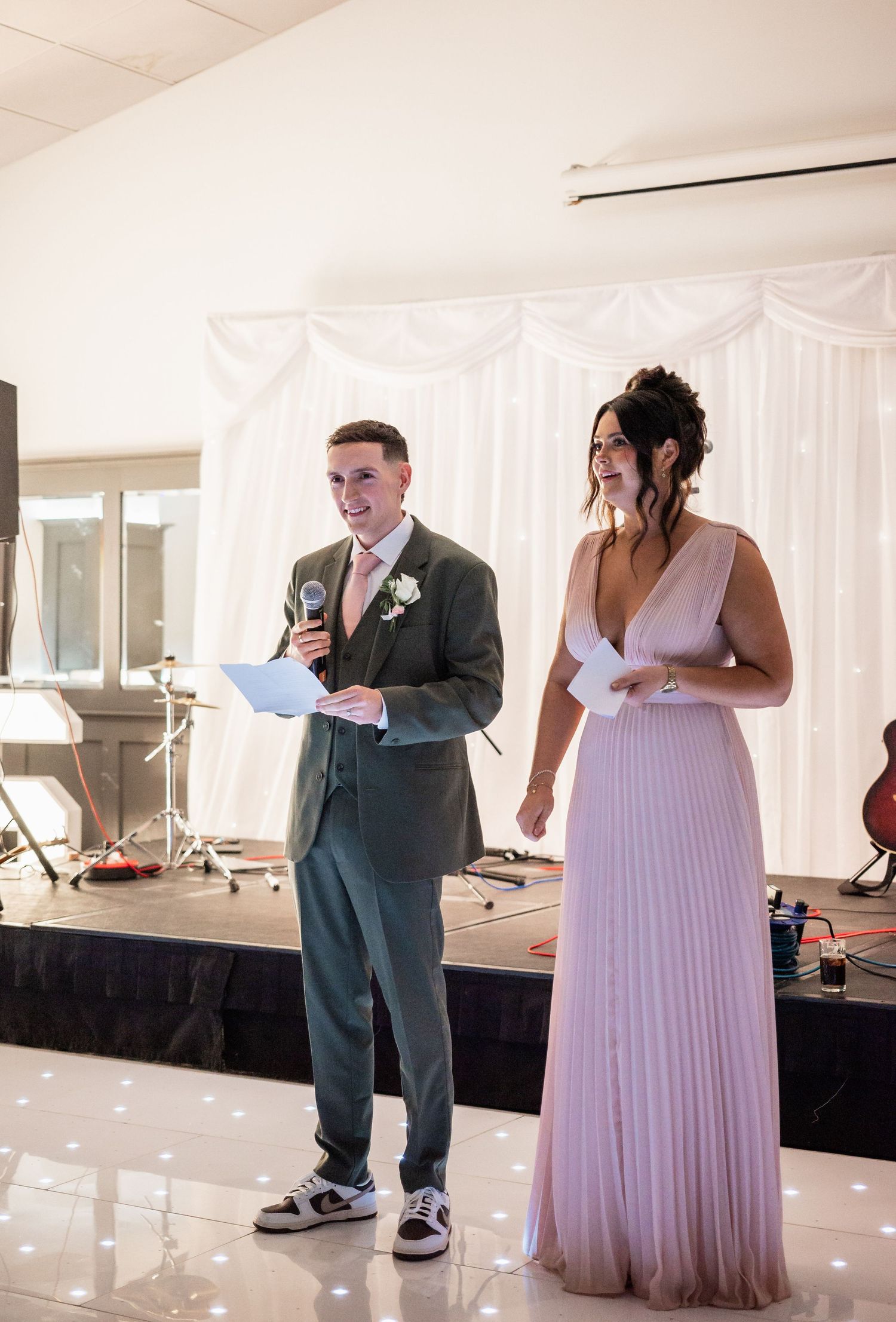 Two people stand on stage during a wedding event with one in gray suit and one in lavender dress.