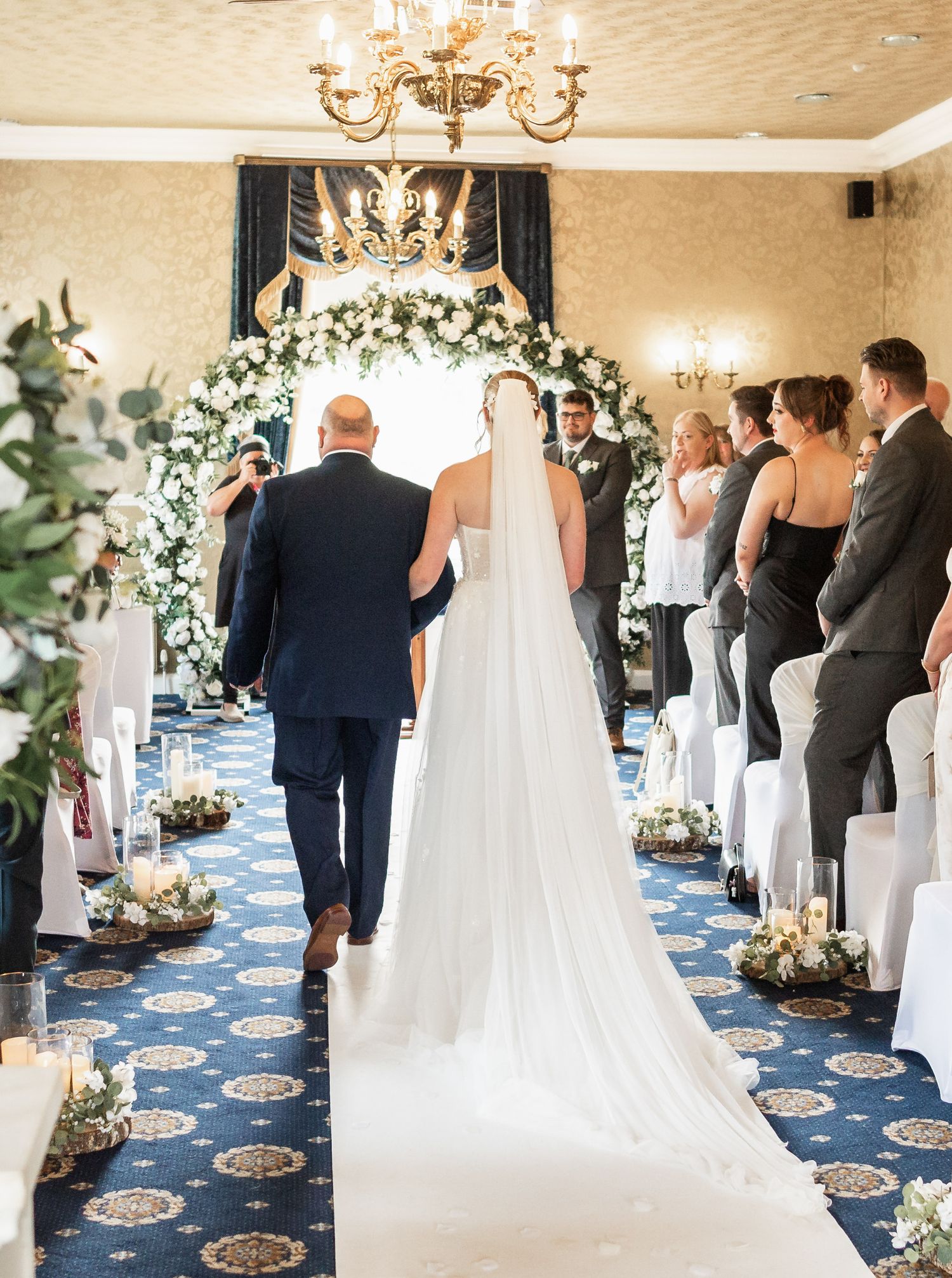 Wedding ceremony in elegant venue with floral arch and chandelier featuring a white aisle runner and ceremony decor.