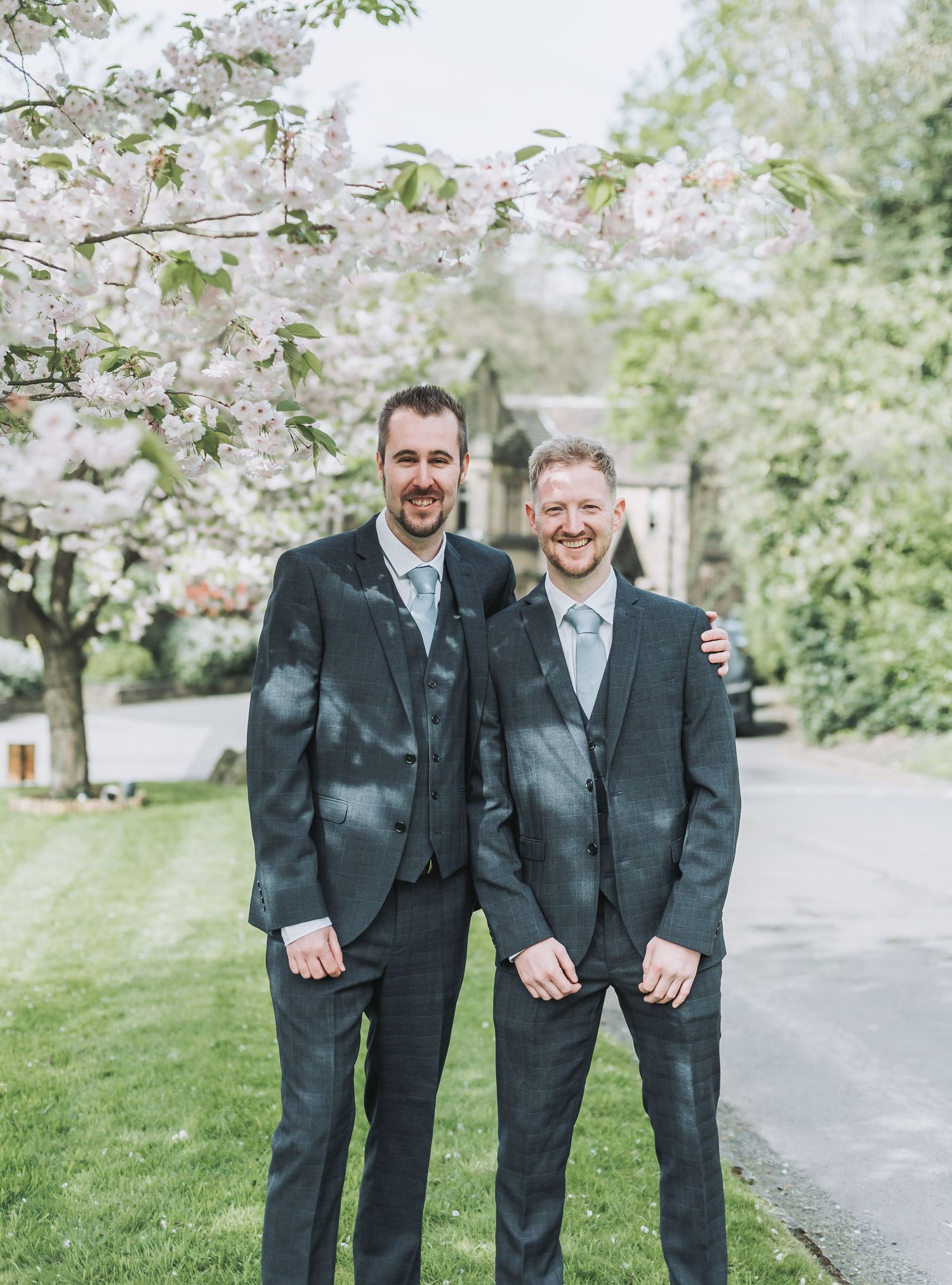 Two grooms in matching dark suits pose under cherry blossoms on their wedding day.