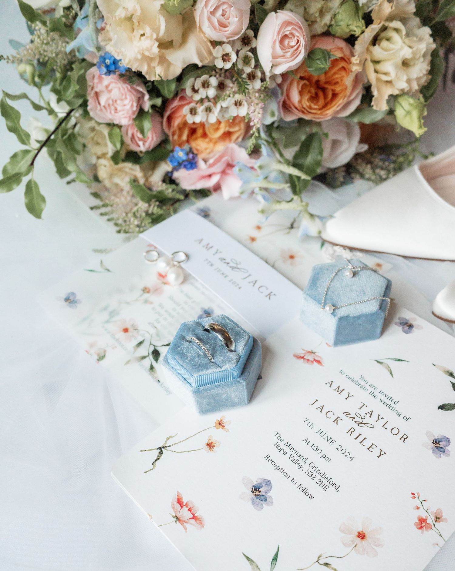 Wedding invitation and blue ring boxes surrounded by delicate floral arrangements.