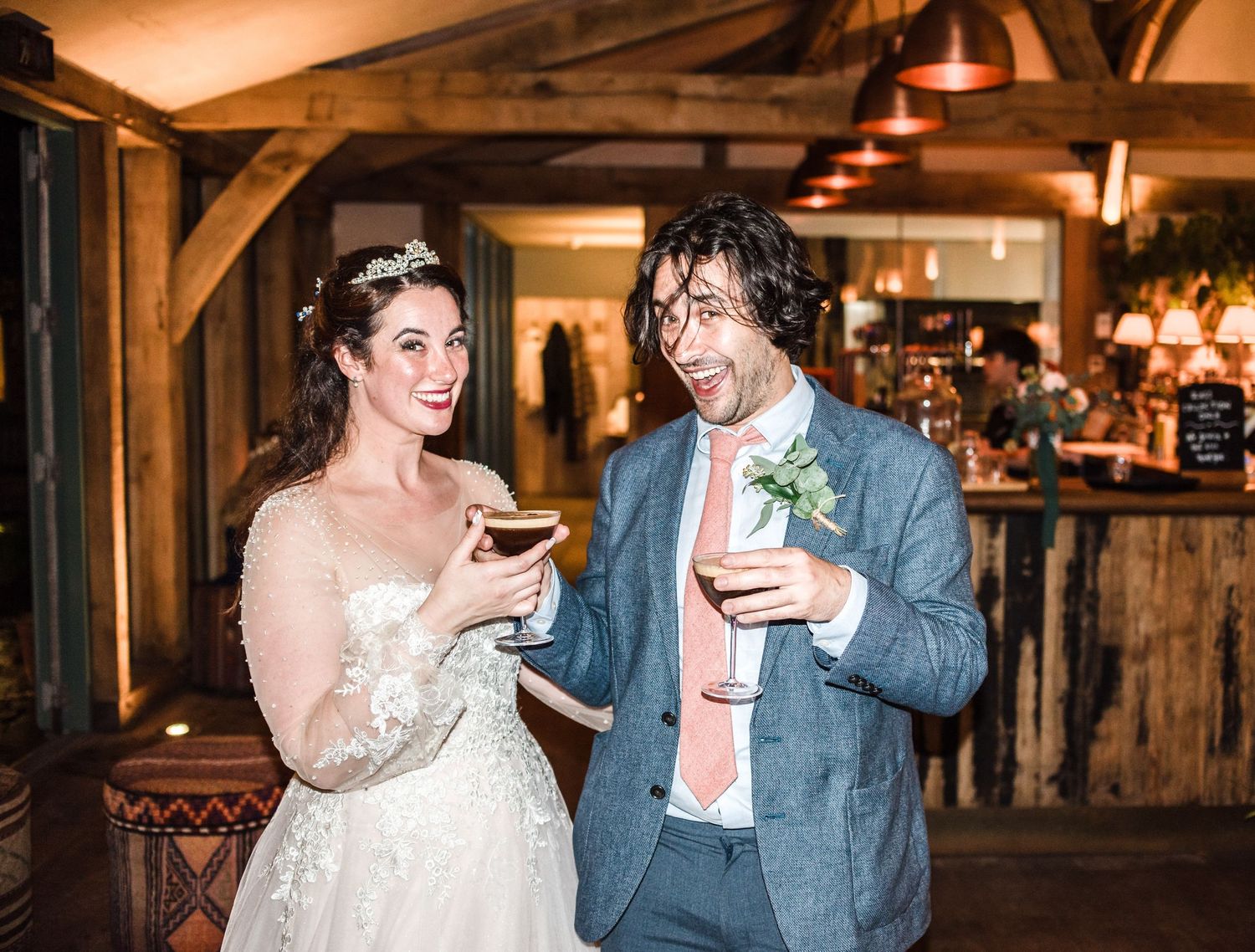 A couple shares drinks and laughter at their reception in a rustic wooden venue with ambient lighting.