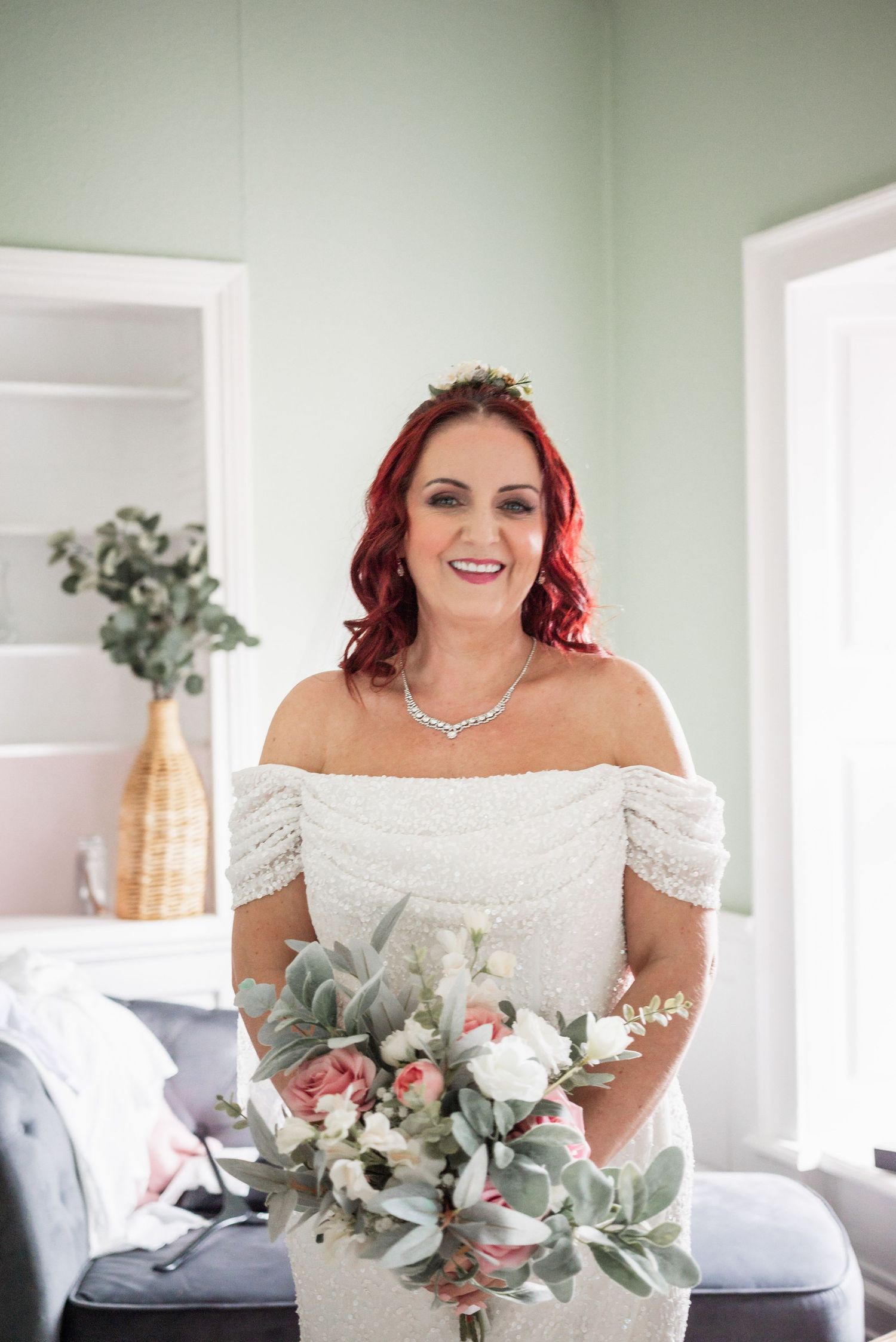 A bride in a white off-shoulder dress holds a bouquet featuring eucalyptus and pale flowers against a light interior.