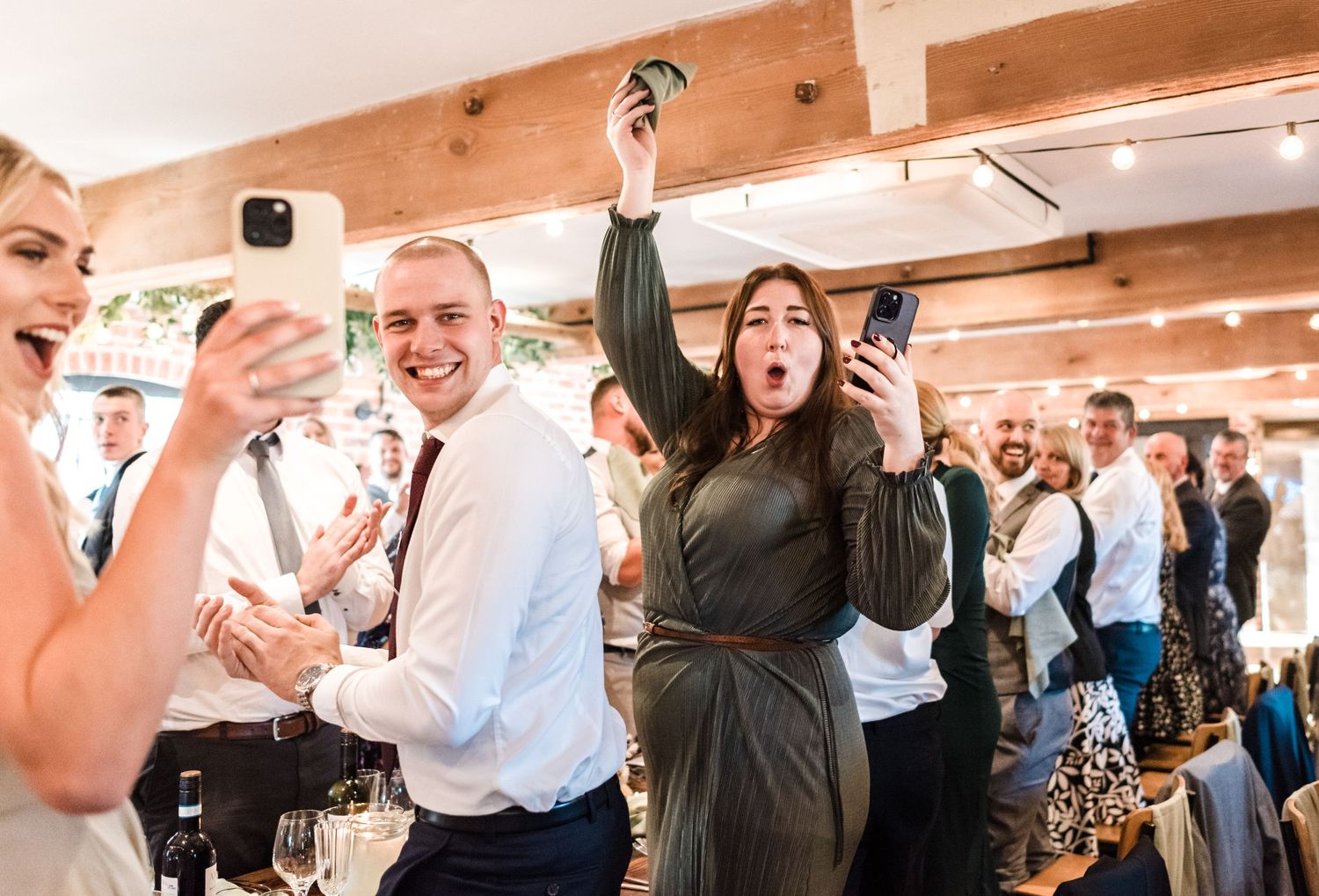 Energetic wedding reception celebration with guests raising glasses in rustic venue.