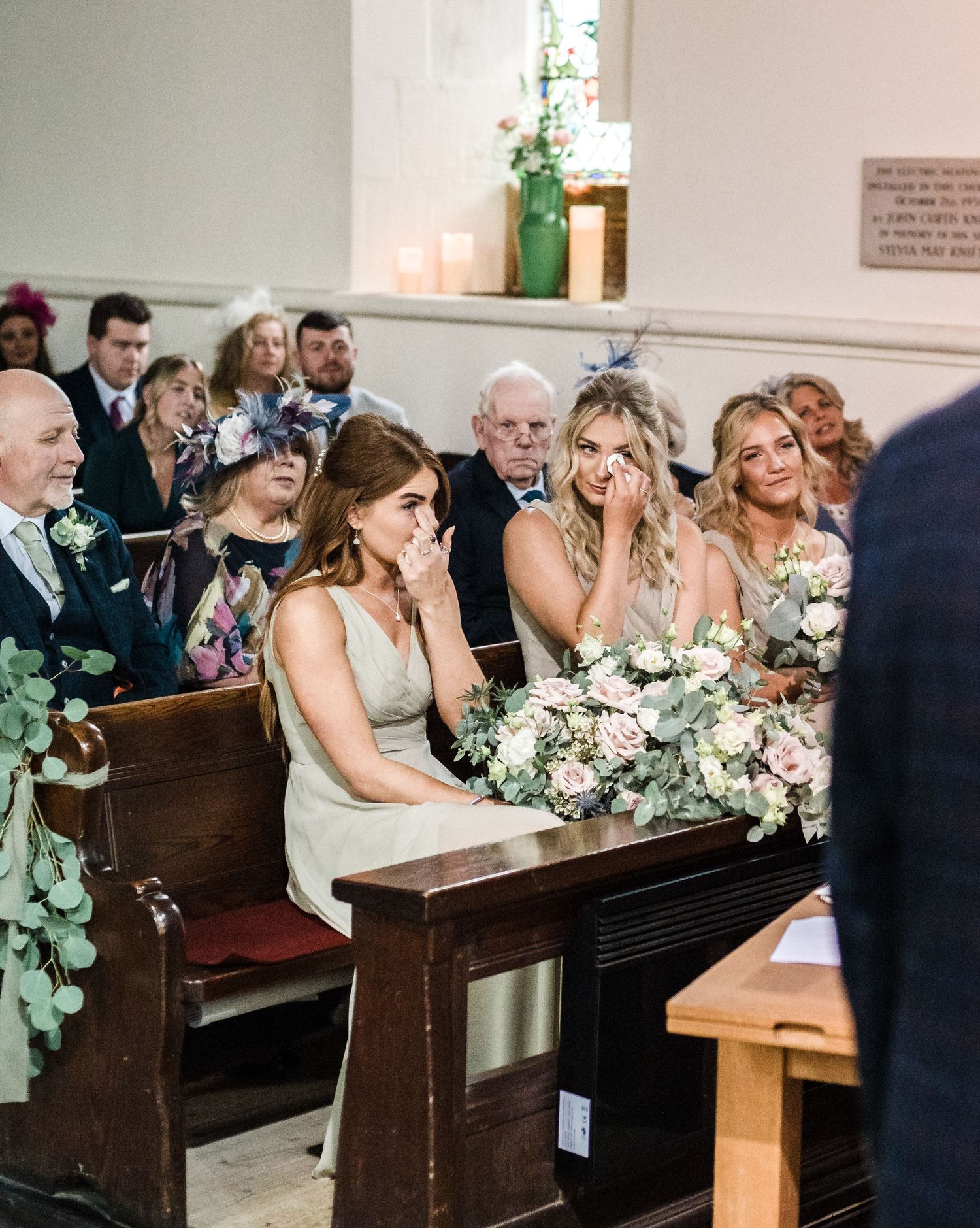Wedding party seated in church pews with elegant floral bouquets and decorative greenery.