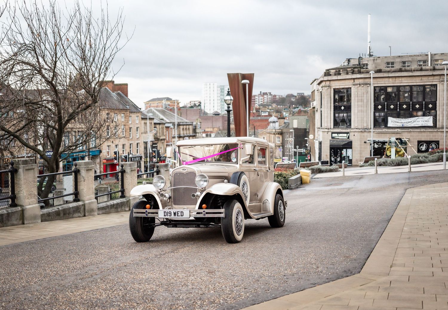 Vintage cream-colored car drives through historic city street on cloudy day.