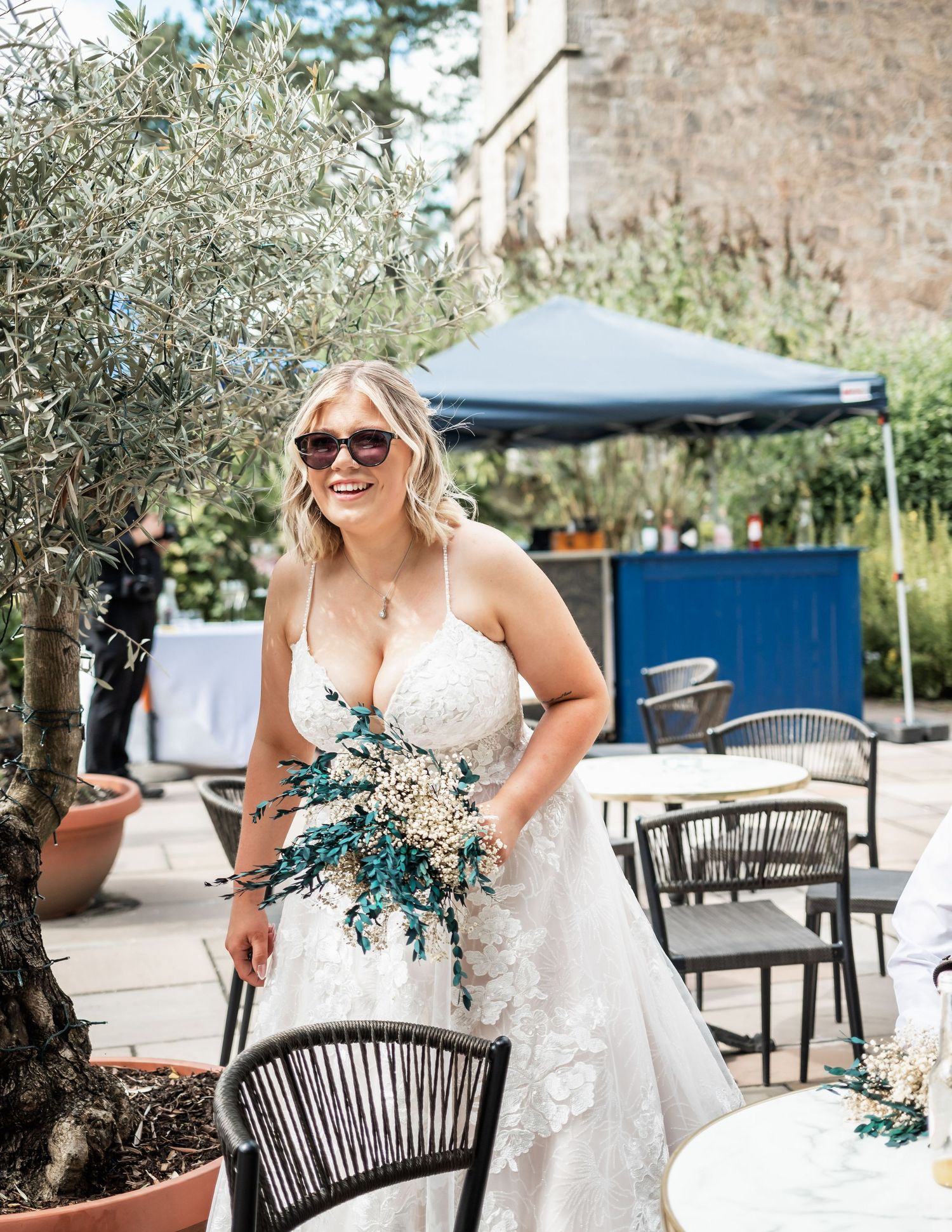Bride in a white lace dress holding a blue and white bouquet poses on an outdoor patio with olive trees.