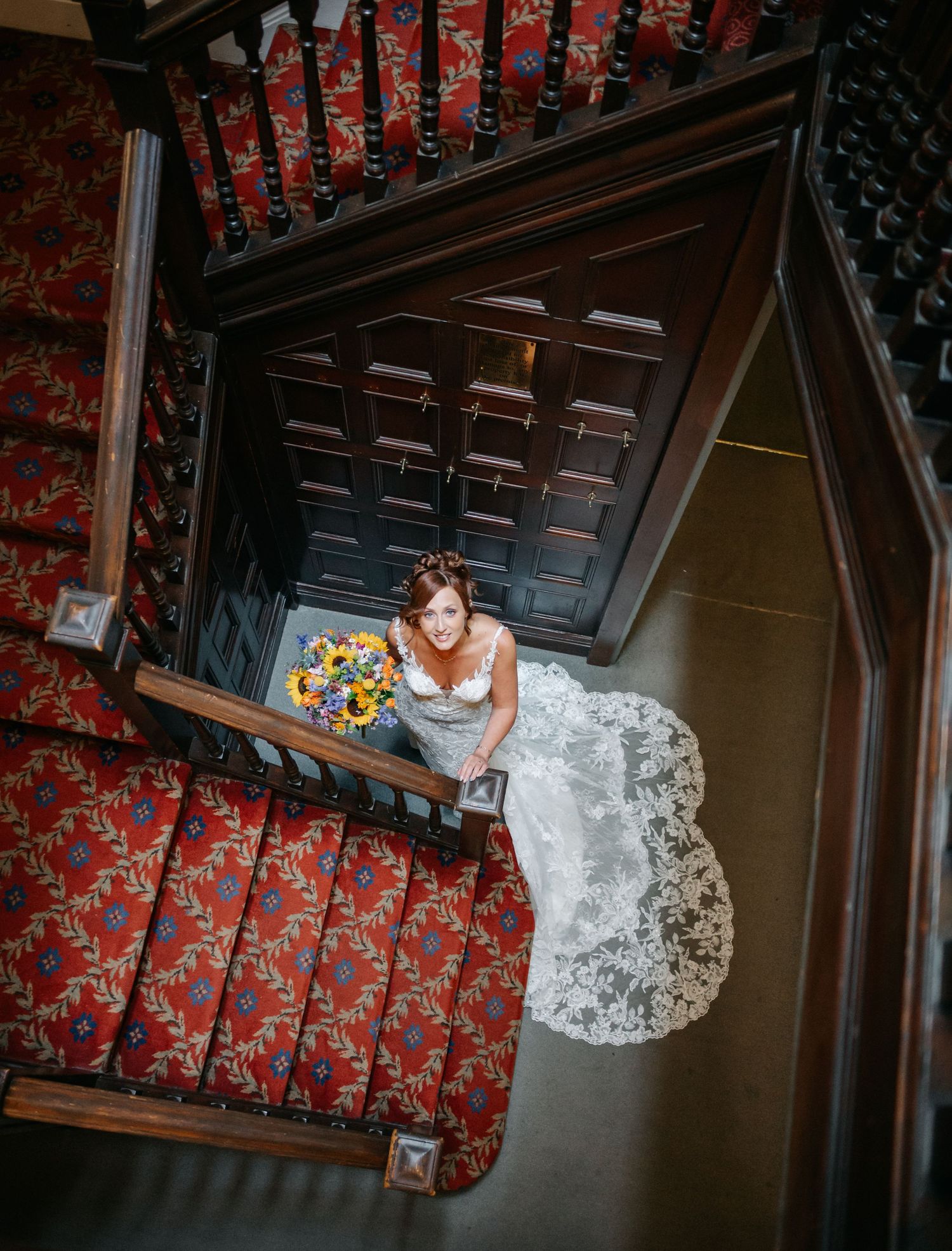 Aerial view of a bride in a white lace dress descending ornate wooden stairs with red carpeting.