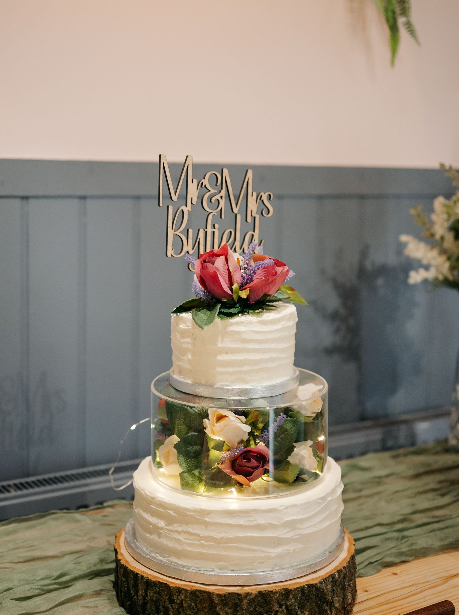 White tiered wedding cake decorated with fresh flowers and 'Mr & Mrs' topper on rustic wood slice.