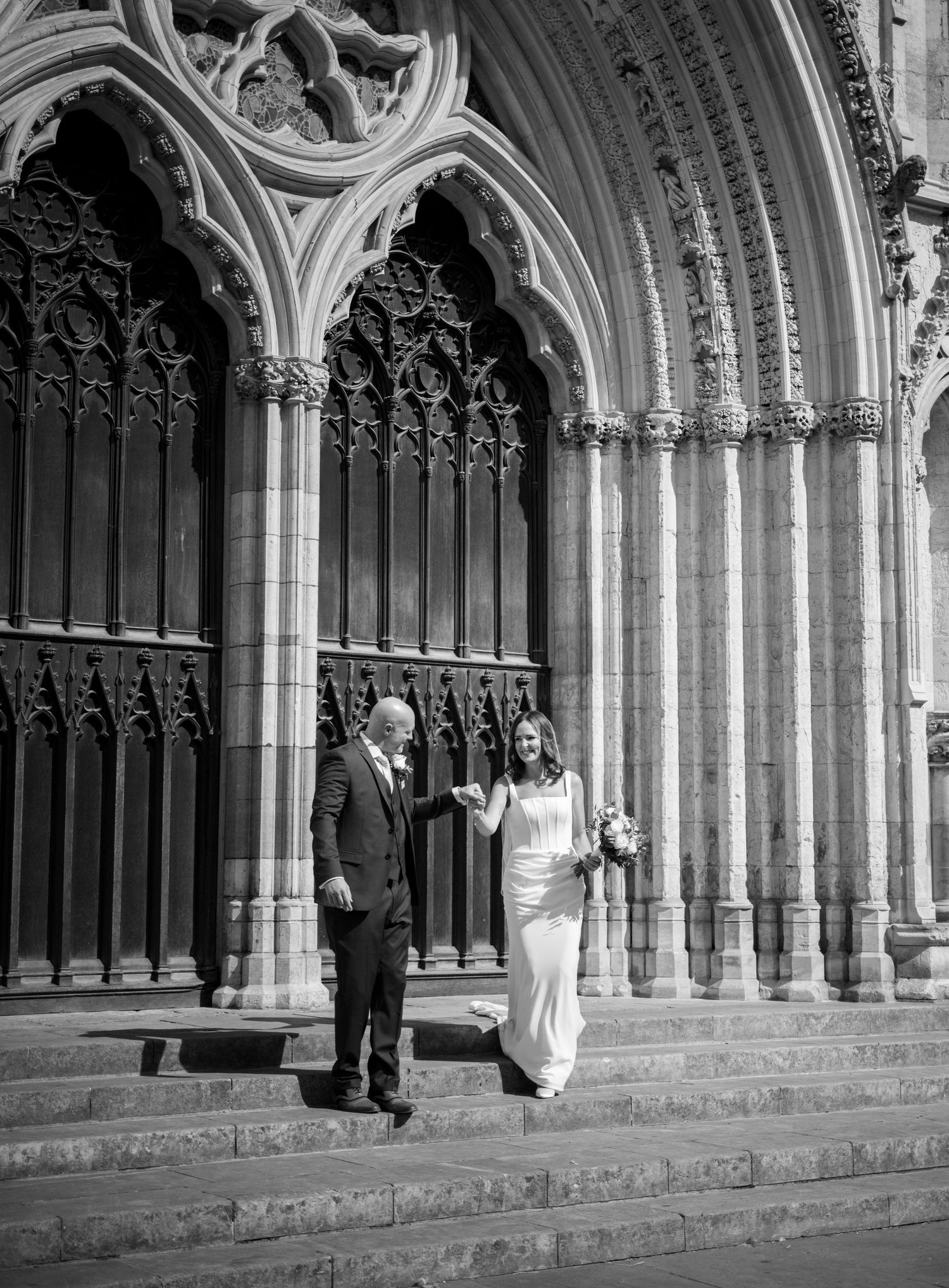 Black and white photo of a bride and groom exiting a grand Gothic cathedral on their wedding day.