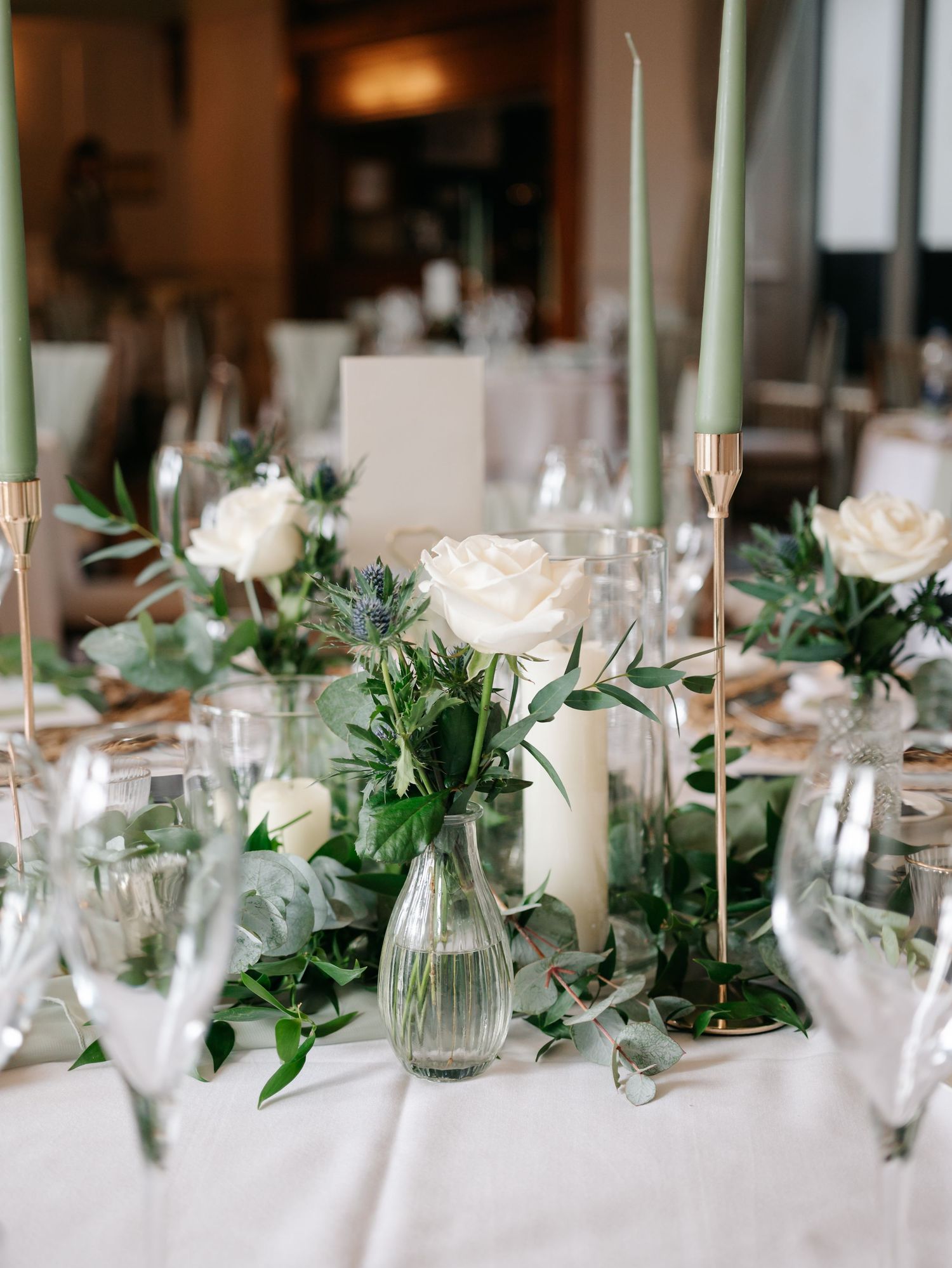 Elegant wedding table setting with white roses, tall green candles, and eucalyptus leaves on a white tablecloth.