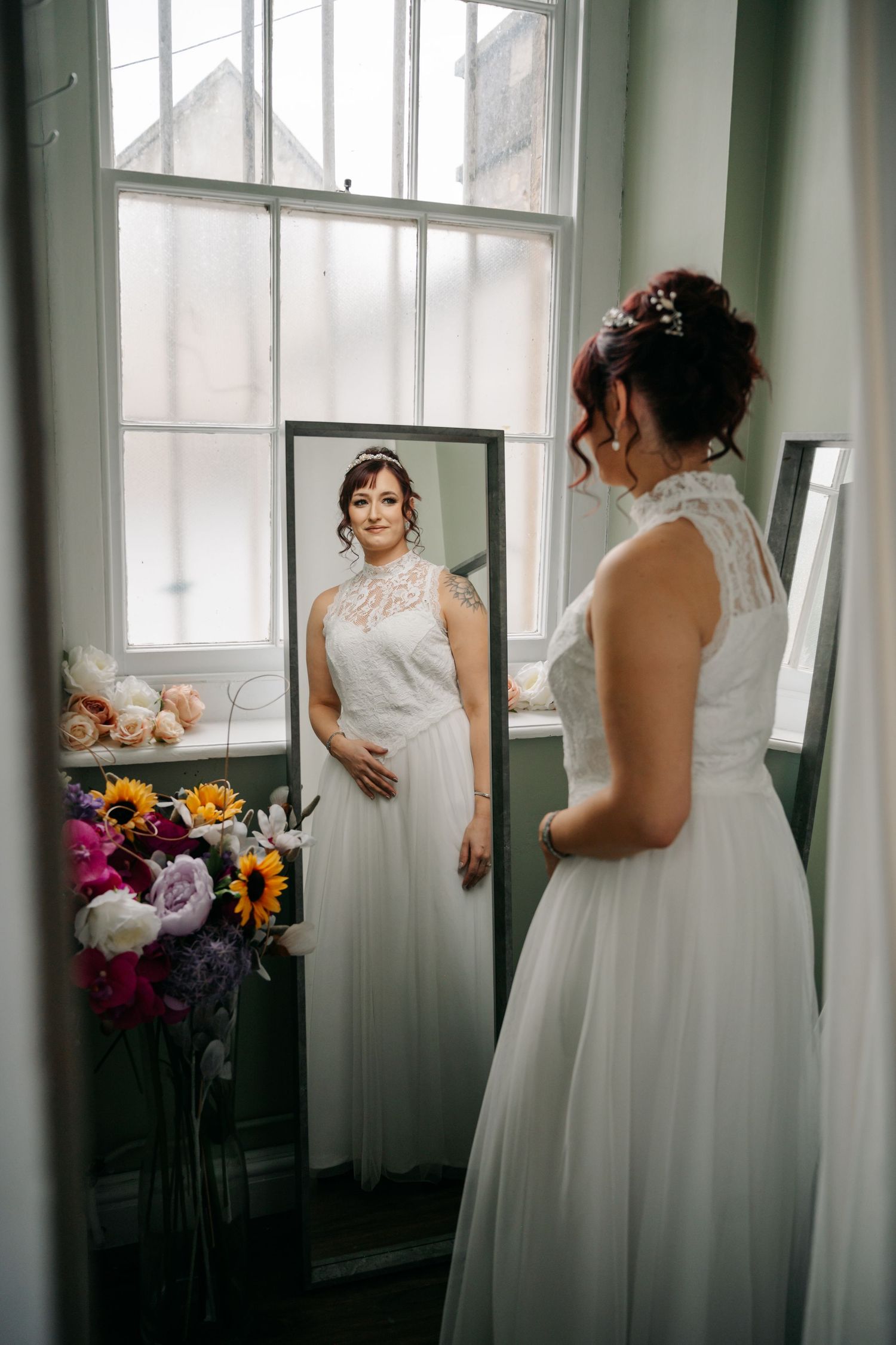 A bride in a white lace dress admires her reflection in a mirror beside colorful floral arrangements.
