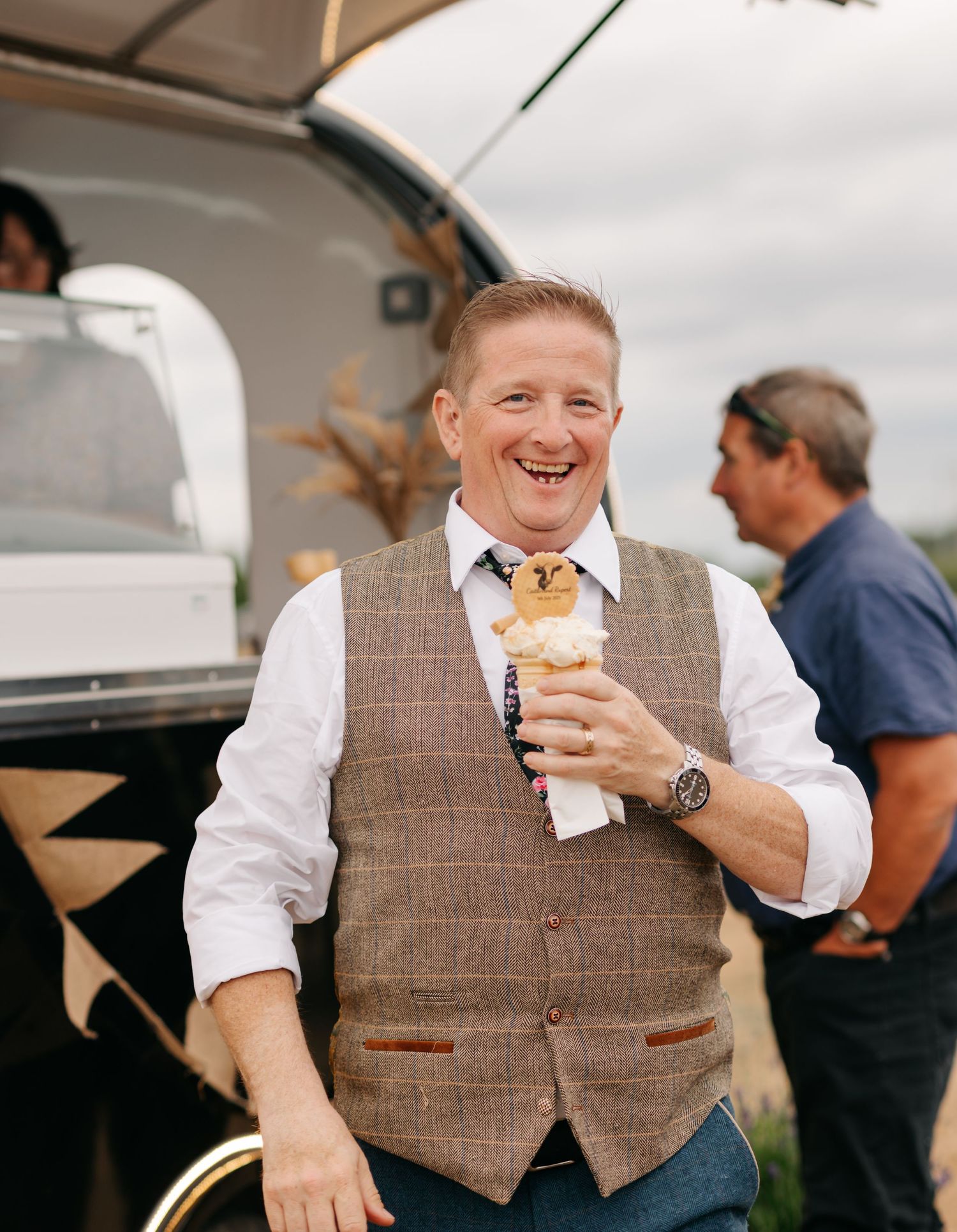 A person wearing a brown tweed vest enjoying ice cream outdoors at a casual event.