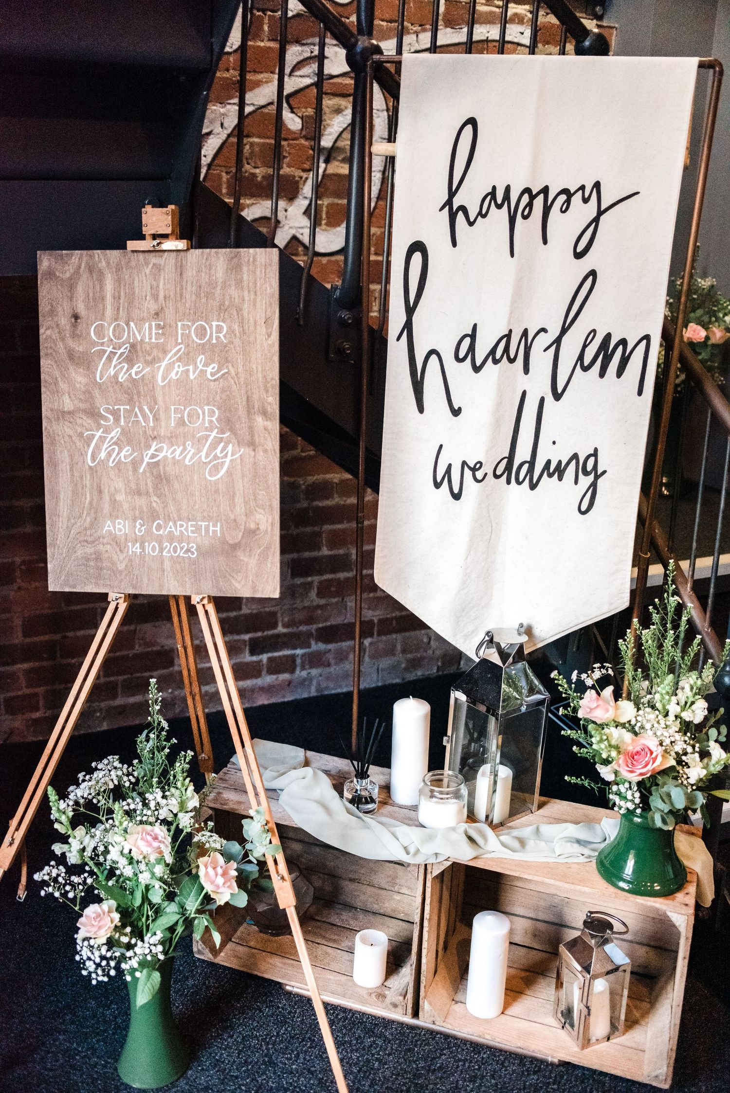 Rustic wedding welcome display with wooden signs, greenery arrangements and candles on a decorative stand.