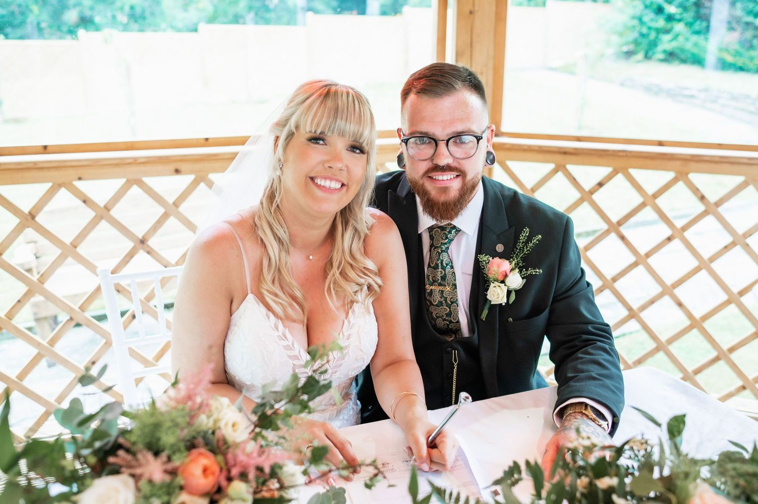 Newlyweds signing documents at wooden gazebo decorated with greenery.