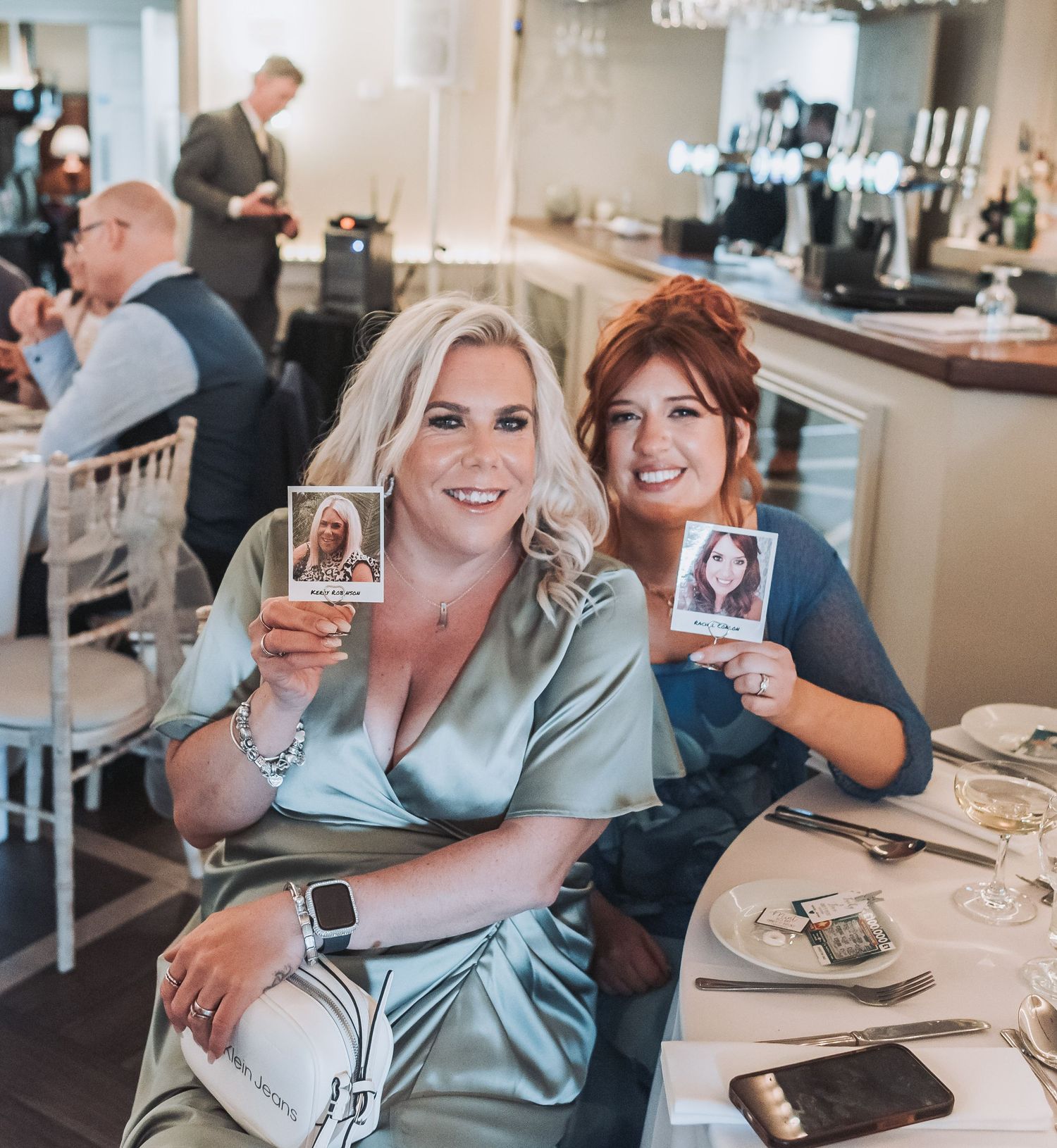 Two friends holding ID cards while sitting at a table during a wedding celebration.