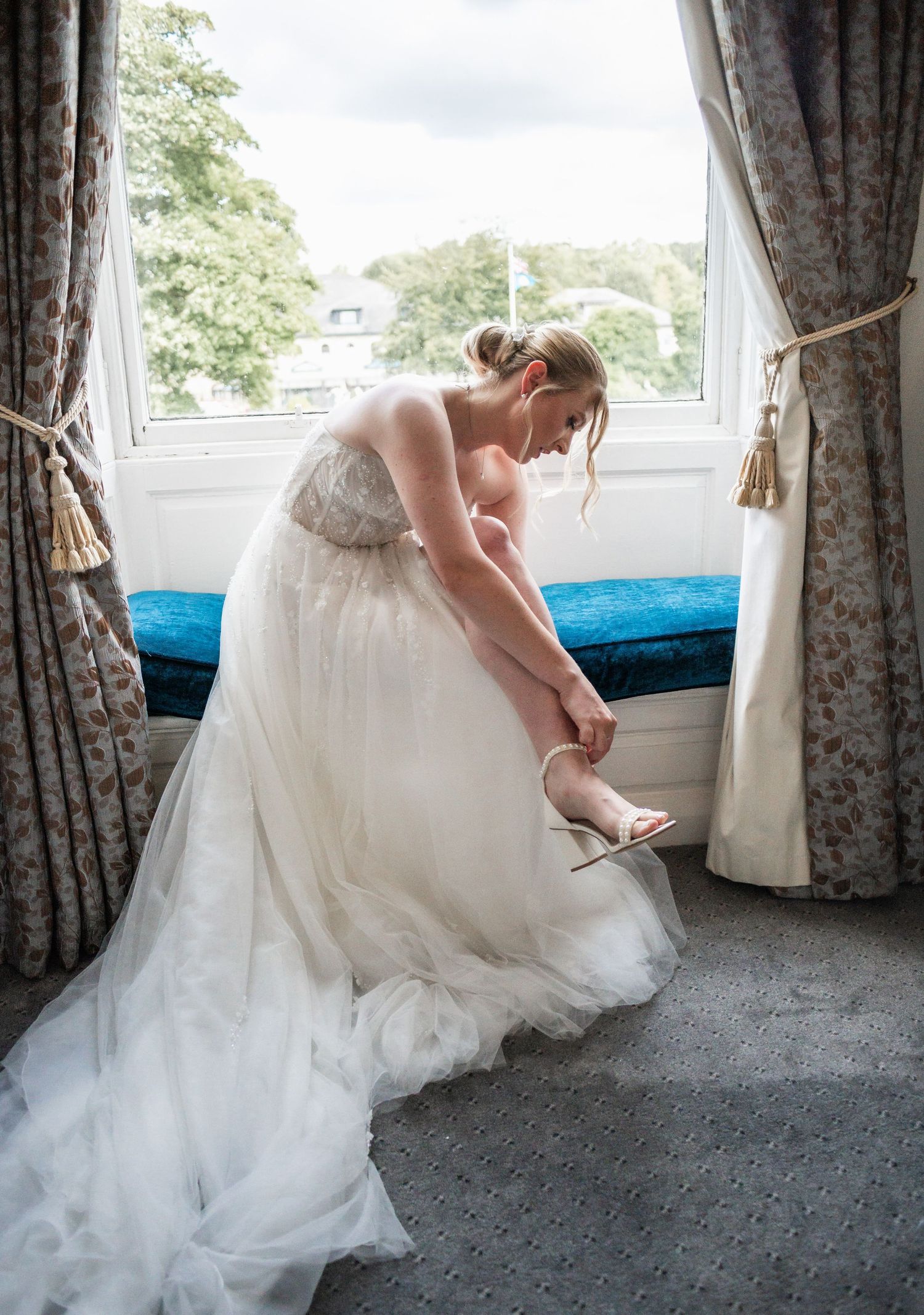 Quiet moment as bride adjusts shoe while sitting by window in flowing white gown.