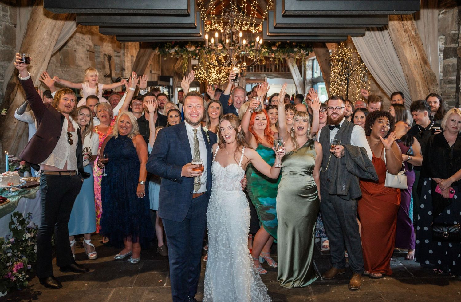 Wedding celebration with guests cheering under elegant chandelier and string lights.