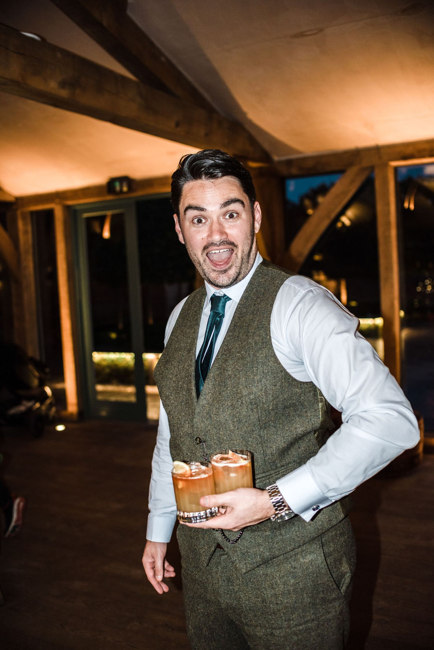 Groomsman in tweed vest poses enthusiastically with cocktails at wedding reception.