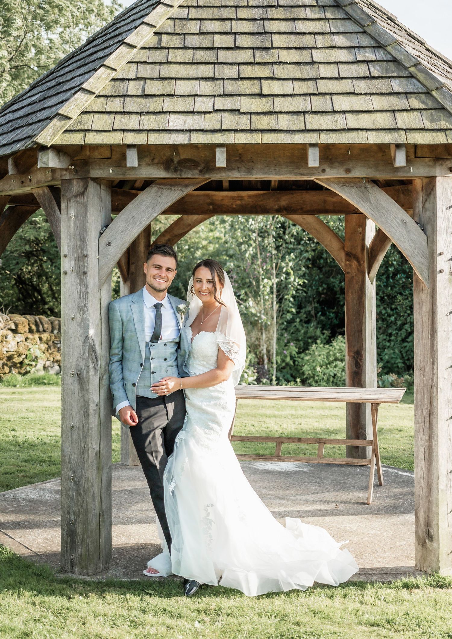 A wedding couple poses under a rustic wooden gazebo with cedar shake roofing in an outdoor garden setting.