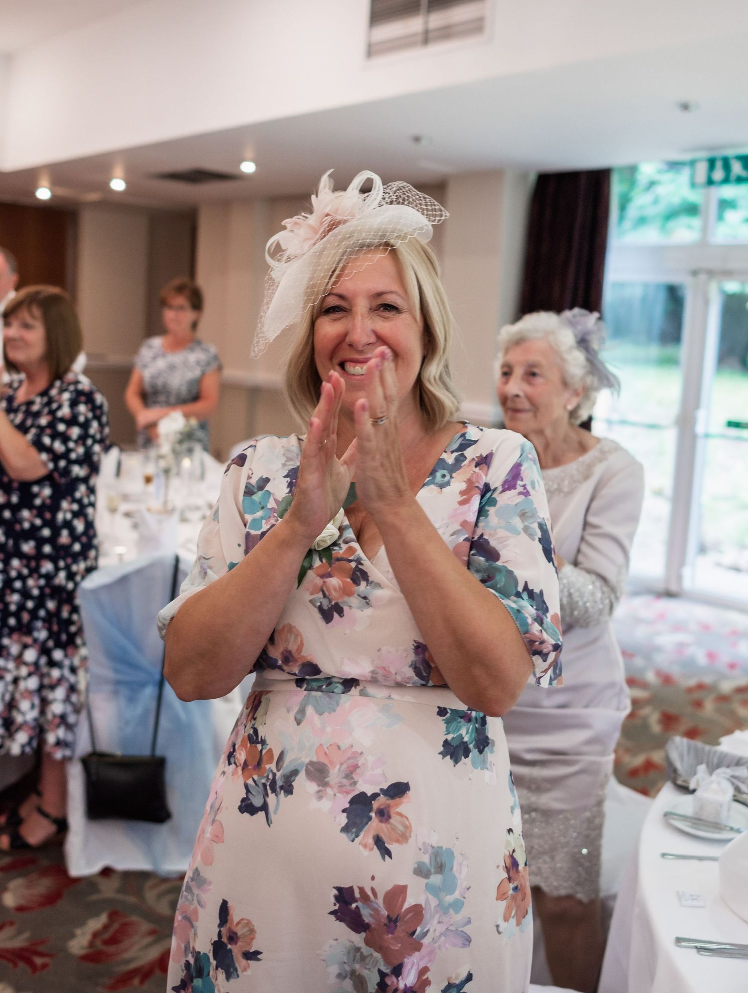 Wedding guest in floral dress claps with joy during reception celebration.