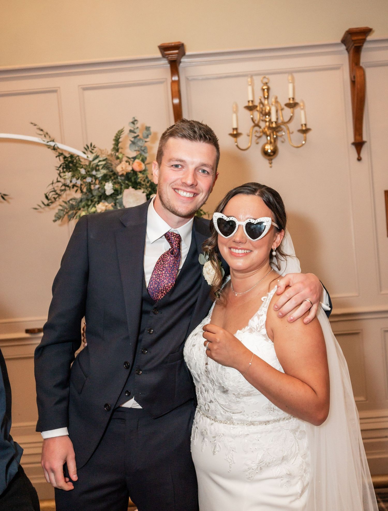 Newlyweds pose playfully with heart-shaped sunglasses next to floral arrangements and candle sconces.