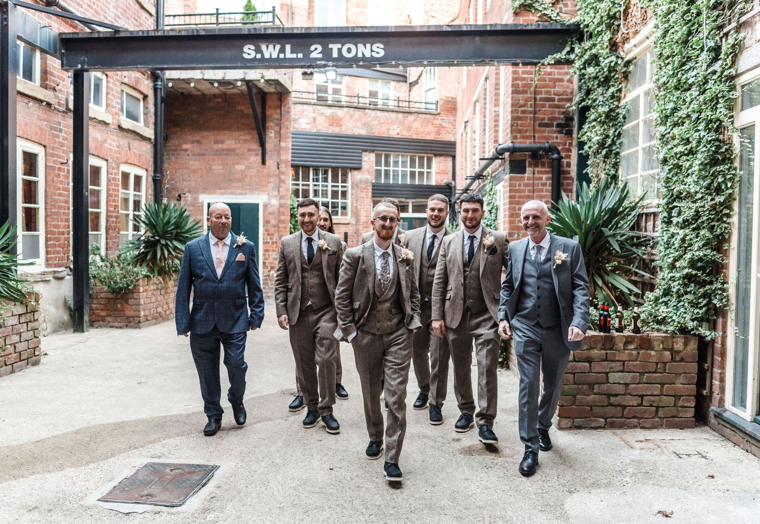 Groomsmen in grey suits walk together outside a rustic brick building.