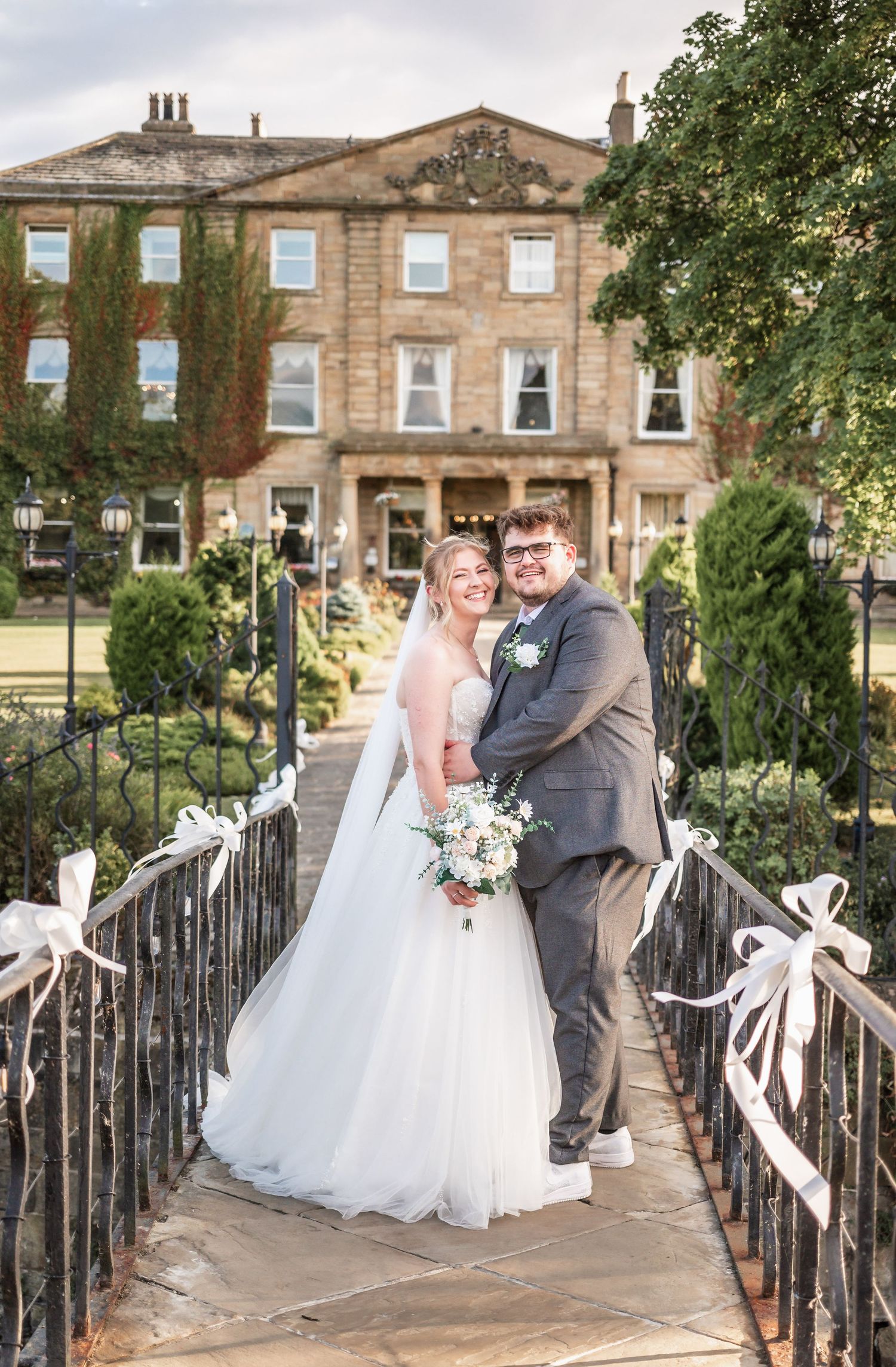 Newlyweds pose on ornate bridge with stunning historic manor house backdrop.