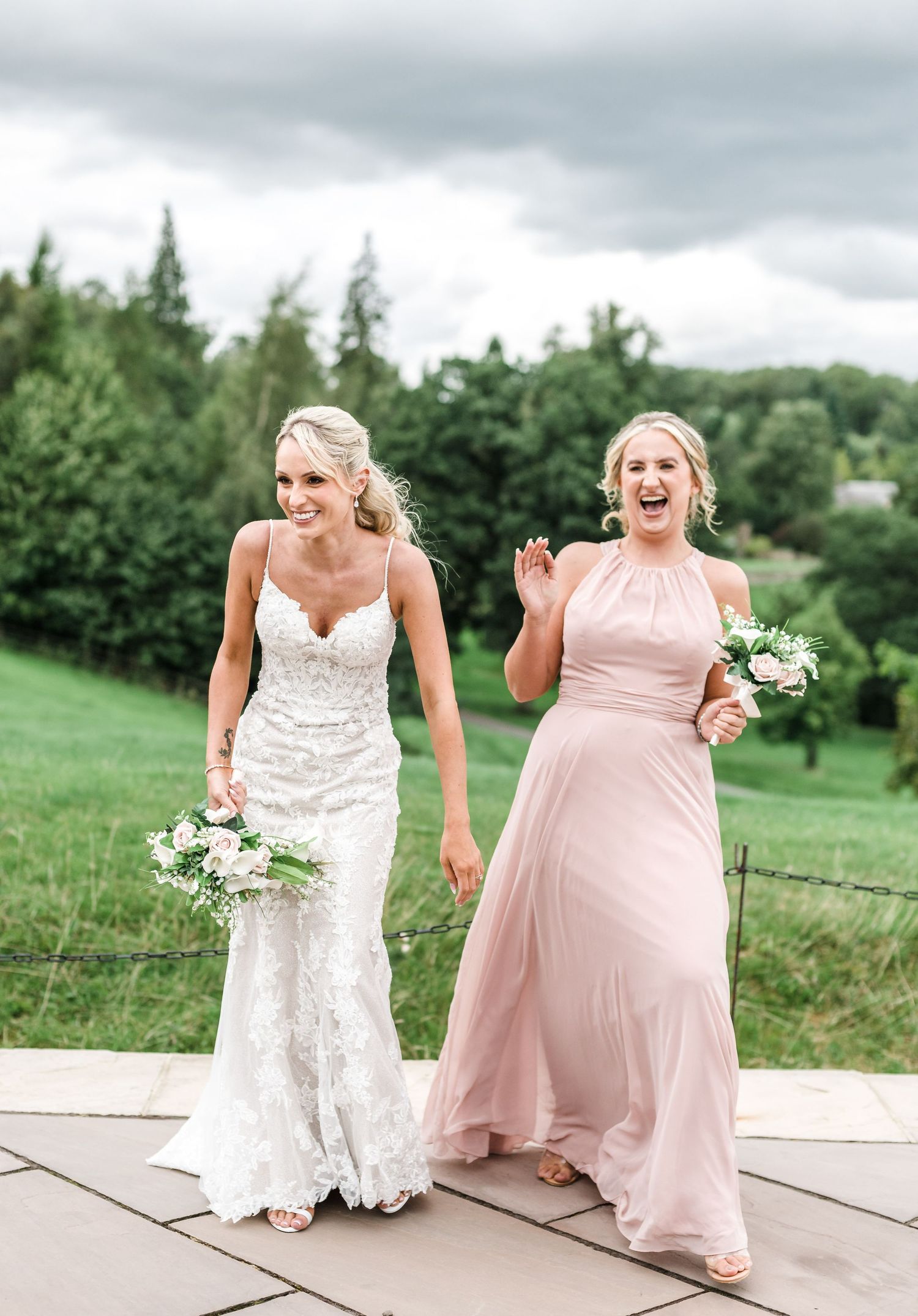 Two women in wedding attire share a joyful moment walking outdoors on a patio surrounded by greenery.