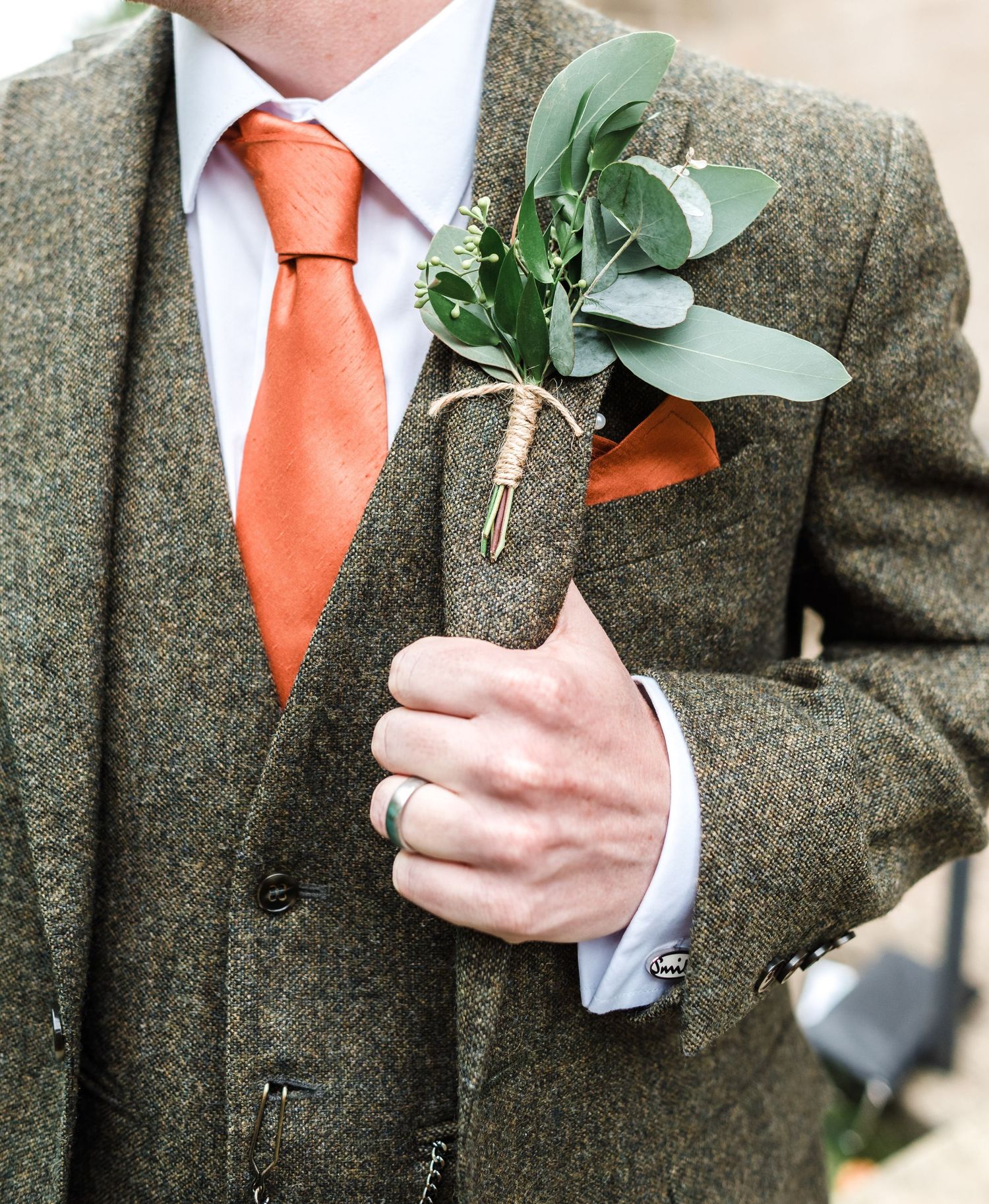 Close-up of groom's boutonniere with eucalyptus on tweed suit with coral tie.