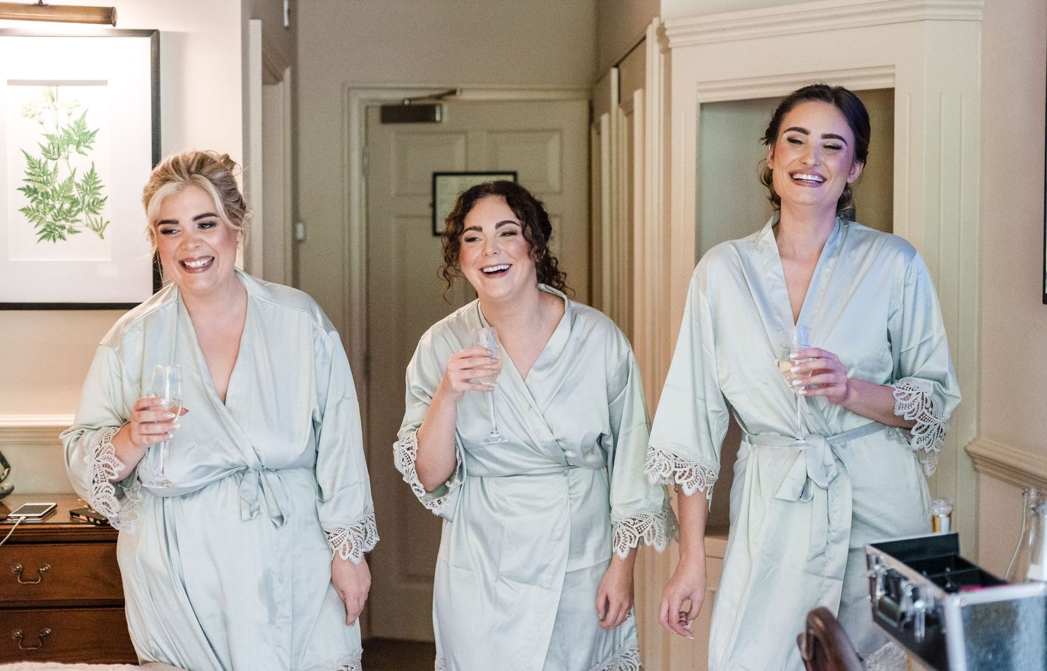 Three women in matching robes share laughs during wedding preparations.