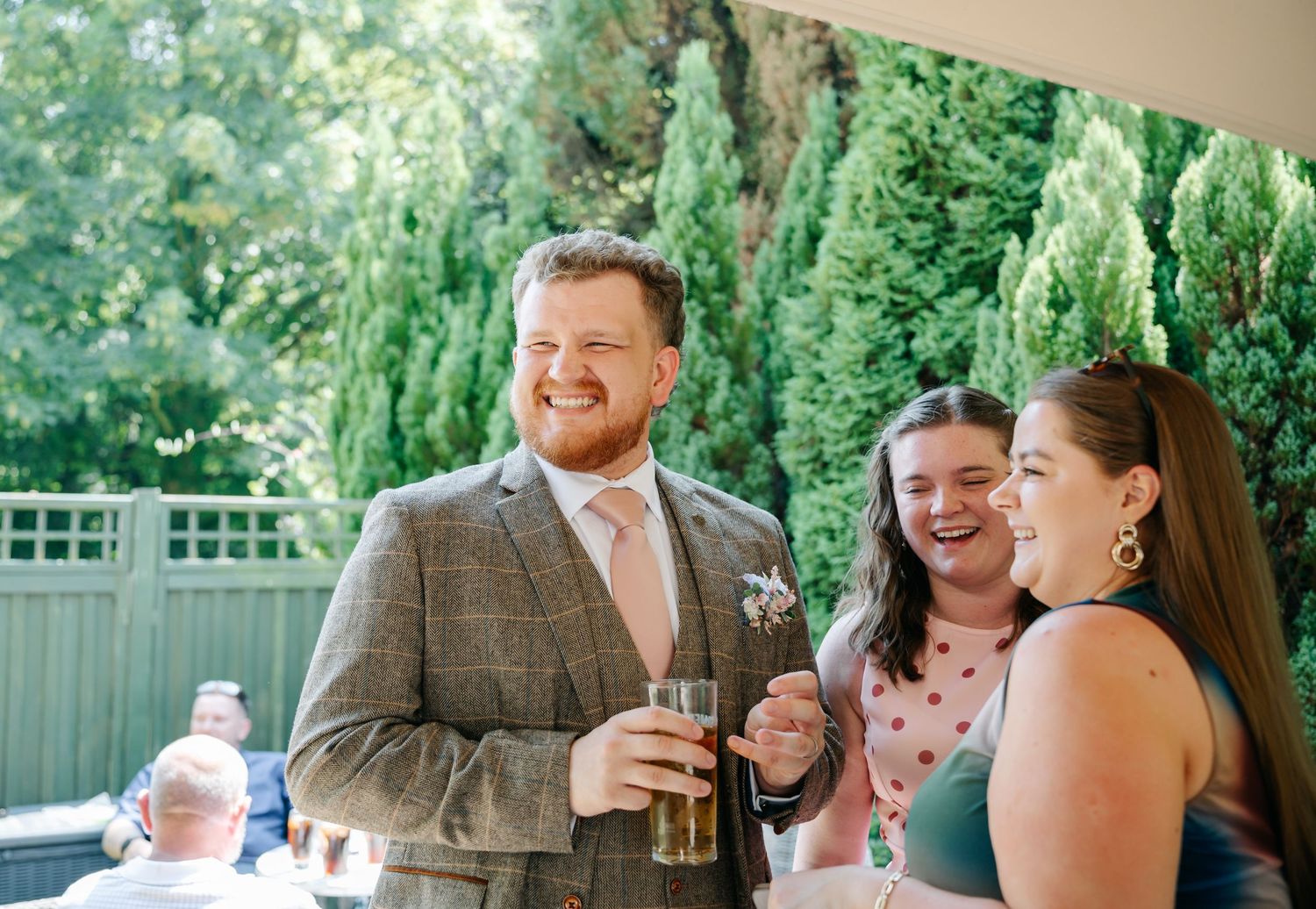 A man in a tweed suit holds a drink while laughing with two women at an outdoor wedding reception.