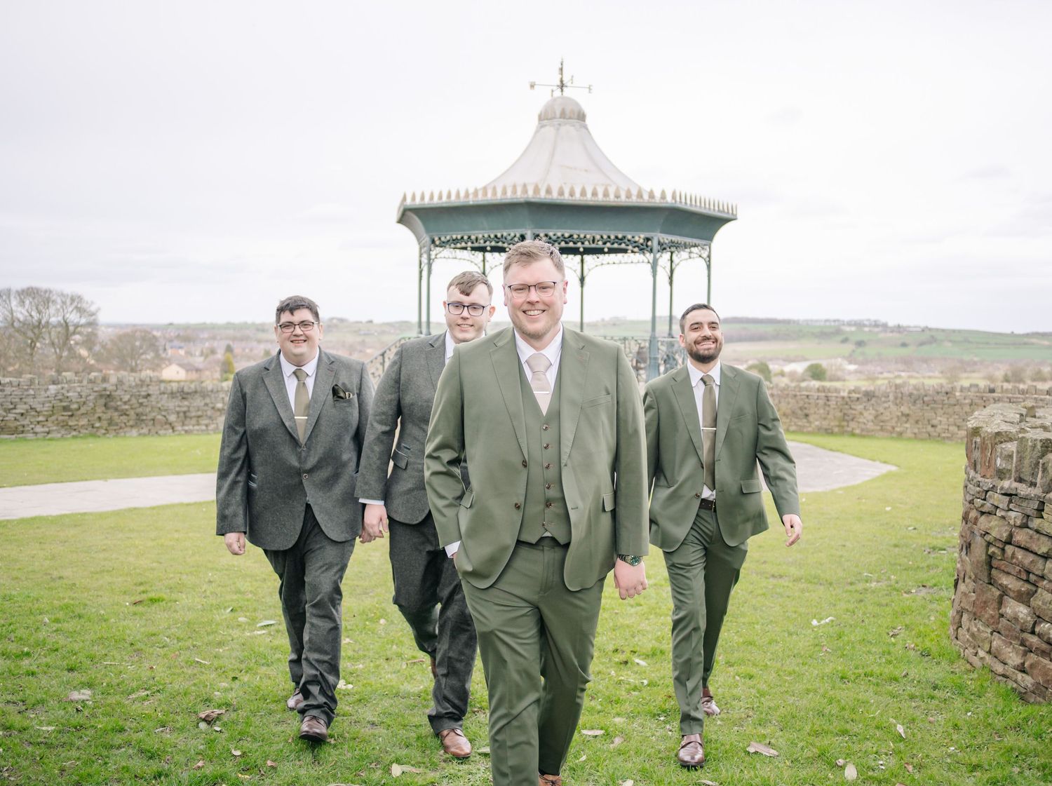 Groomsmen in dark suits walk along a scenic outdoor path with stone walls and green fields in the background.