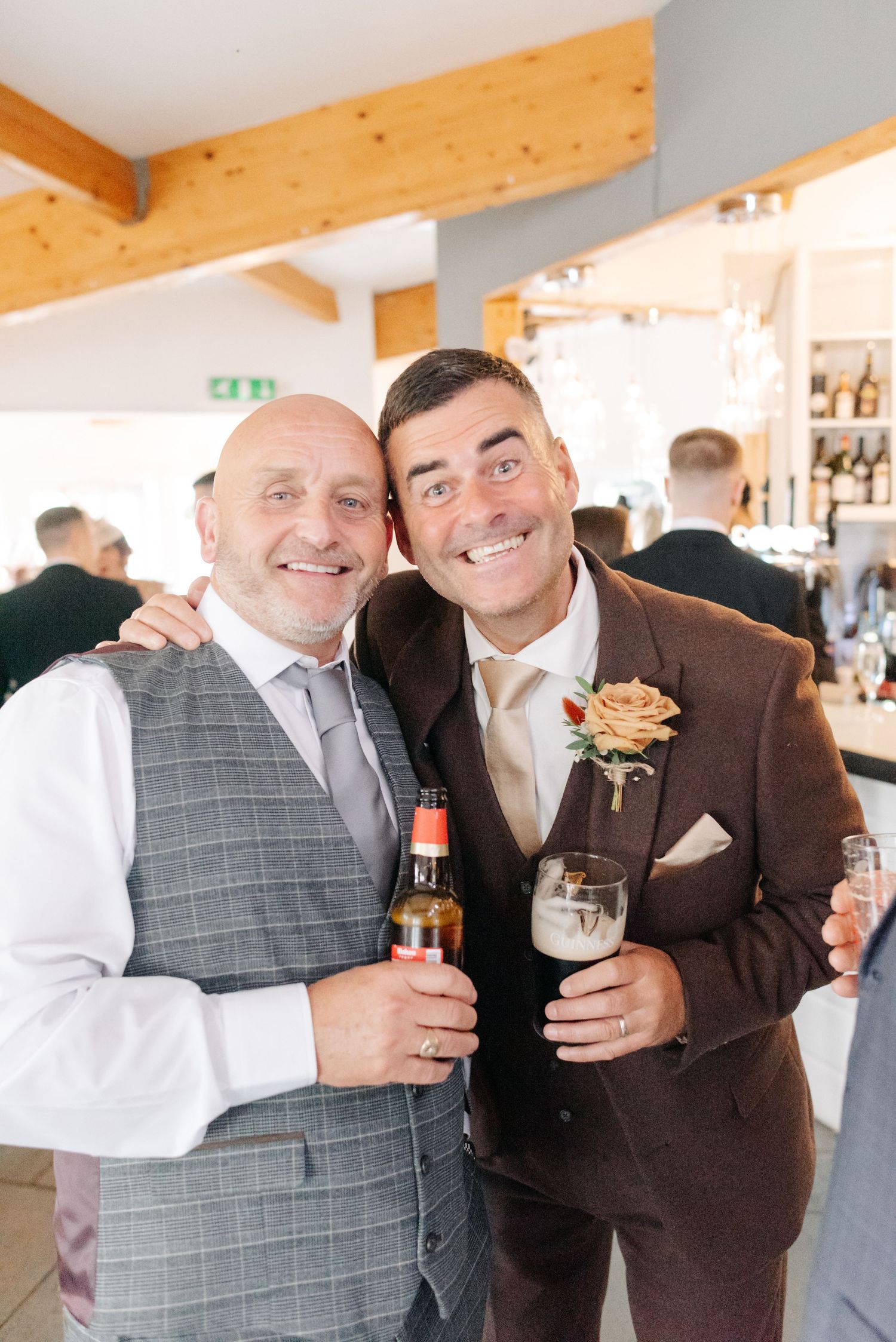 Wedding guests in formal attire sharing drinks and laughs at an indoor reception venue with wood beam ceiling.