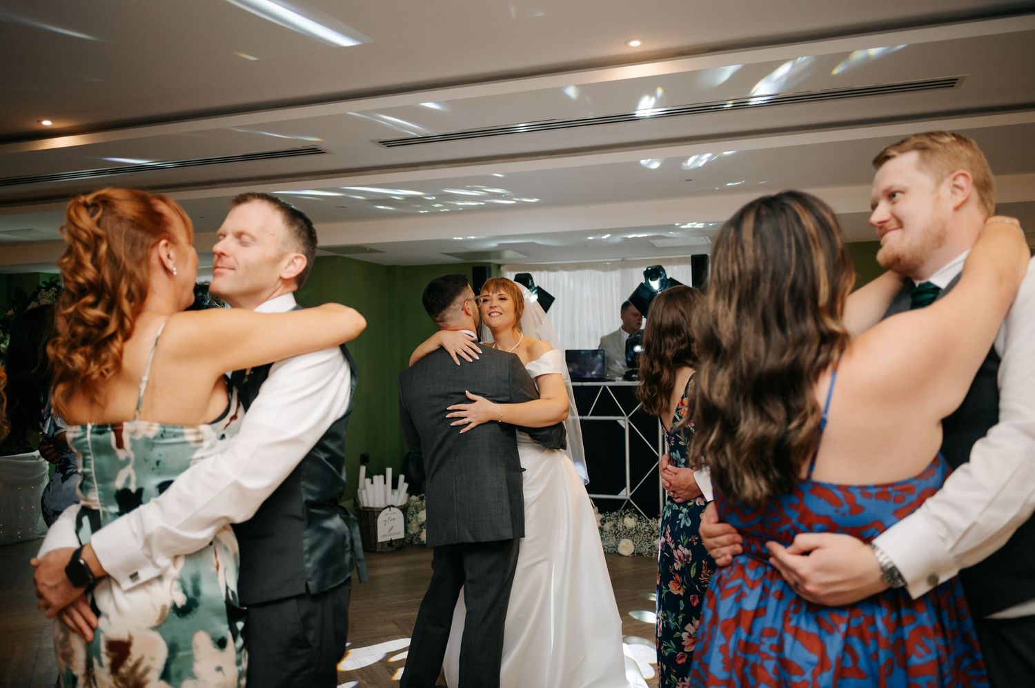 Wedding celebration with guests hugging and dancing in an indoor venue.
