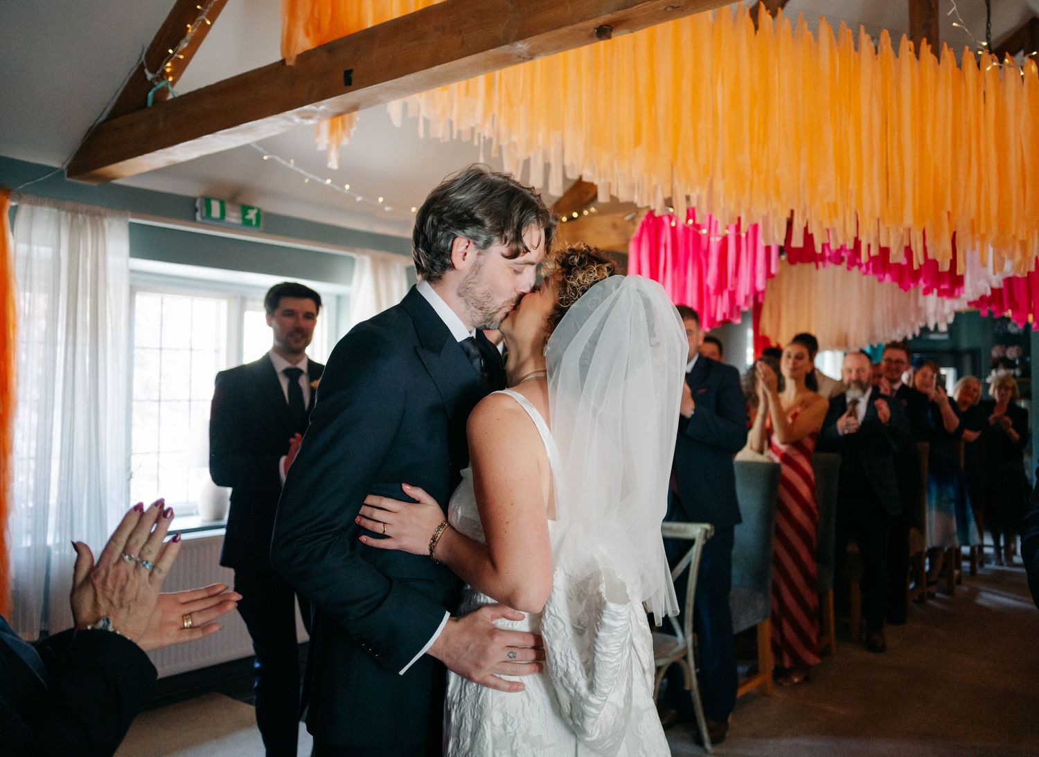 A romantic first kiss moment during a wedding ceremony under colorful streamers.