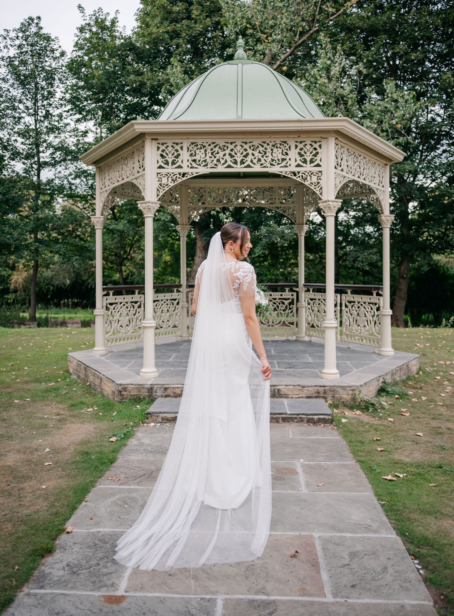 A bride in a flowing white gown twirls and poses in an ornate garden gazebo during a wedding photoshoot.